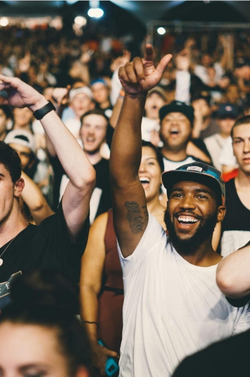 A diverse group of trade professionals at an event. A man in the foreground is raising his hand with a thumb up and making an 'L' shape with his fingers, wearing a white t-shirt and a baseball cap. Market to trade pros in masses.