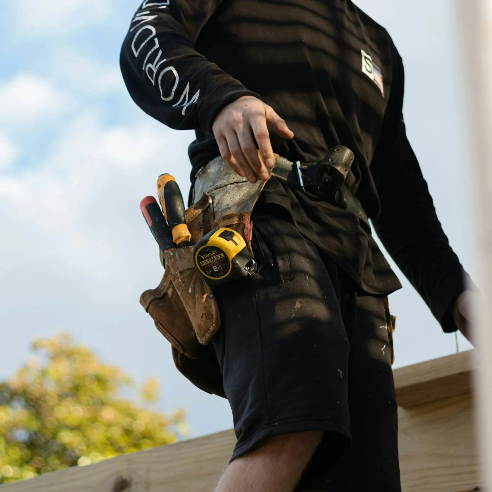 Construction worker wearing black clothing with a tool belt containing screwdrivers, a measuring tape, and a hammer, working outdoors. Know more about your trade pros. Customer data.
