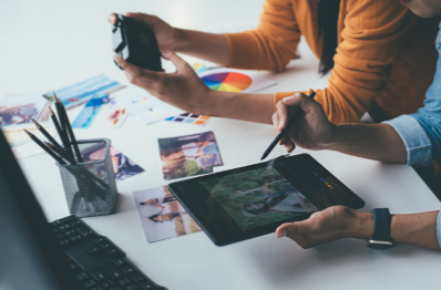 Desk with color swatches, a drawing tablet displaying color diagrams, a ipad, people working on creative ideas, and various paper samples indicating creative concepting.