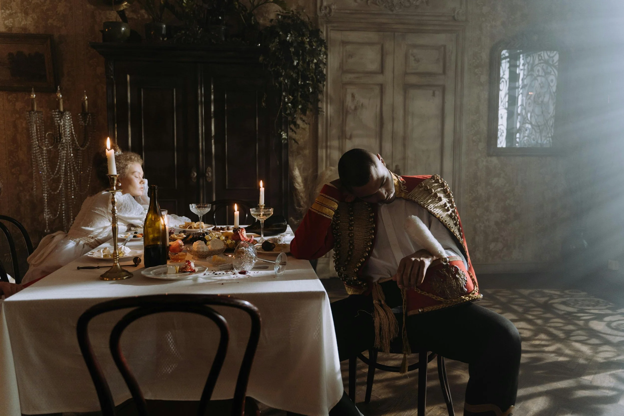 A man and a young girl are seated at a dining table with food and candles, in a dimly lit room, with the man dressed in a Spanish matador costume and the girl in period costume.