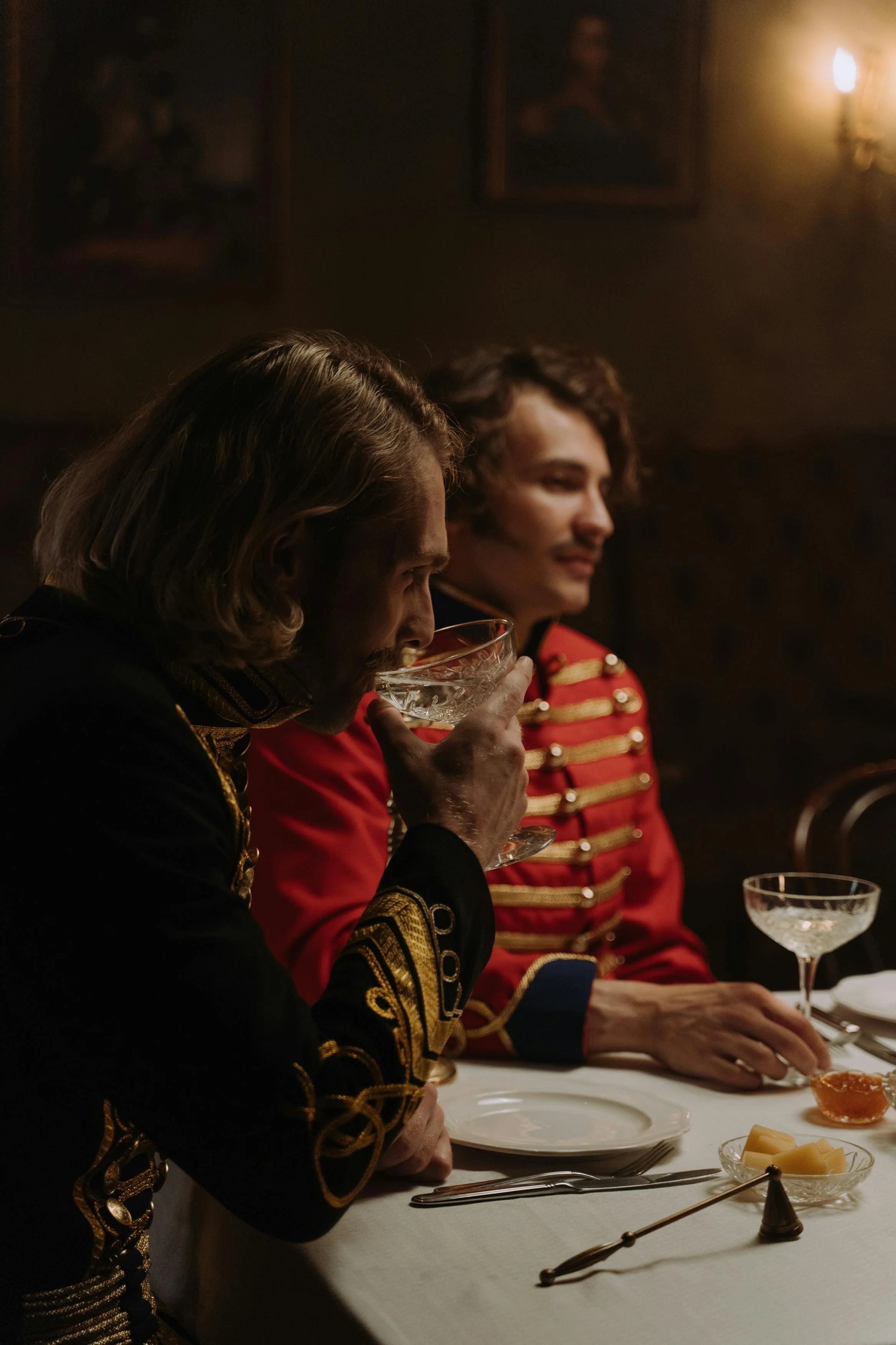 Two men in historical military uniforms sitting at a dinner table, one drinking from a glass, in a dimly lit room with paintings on the wall.