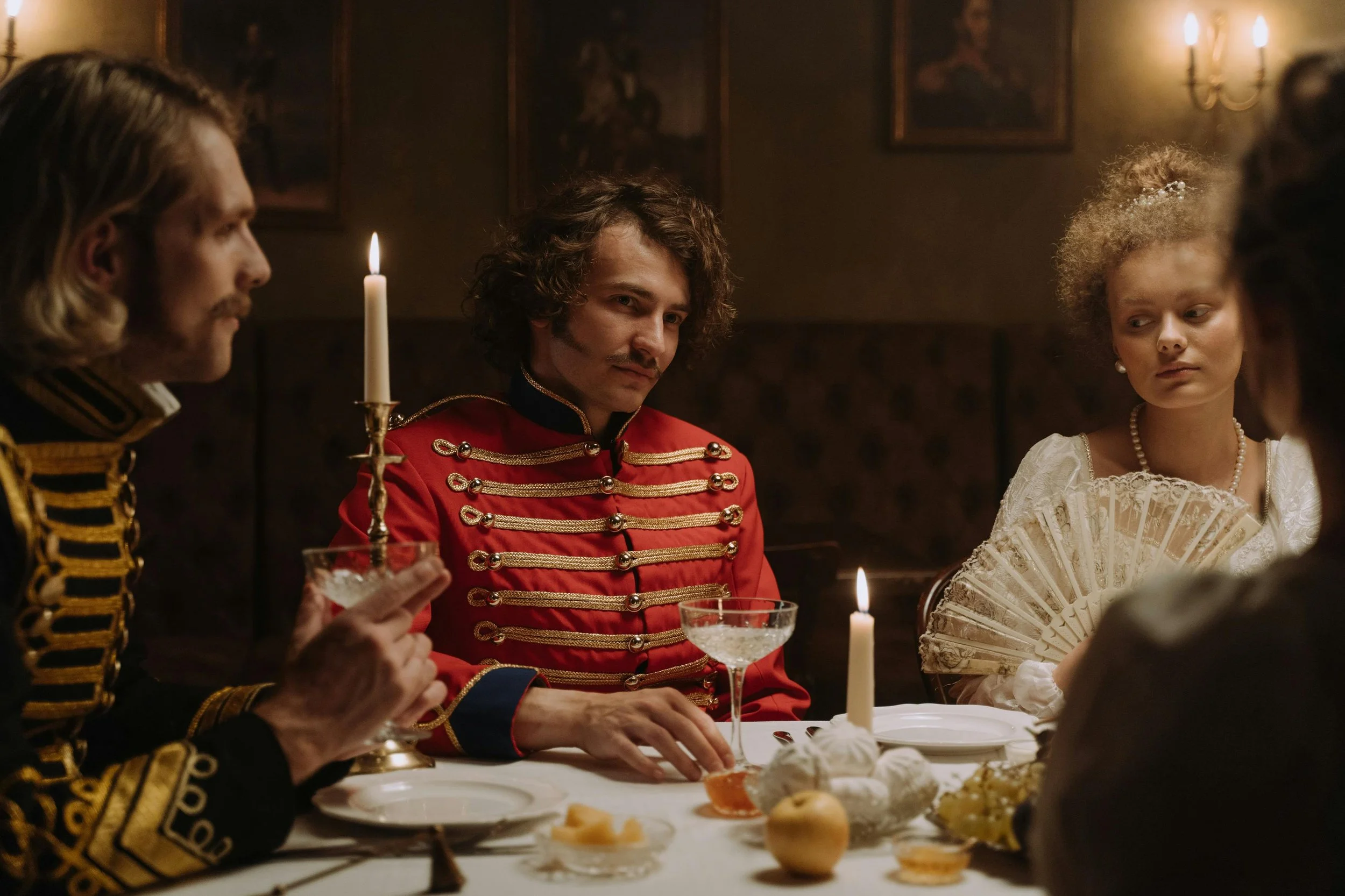 People in elegant historical costumes sitting at a candlelit dinner table, with a man in a red military-style jacket at the center, holding a glass, in a vintage-style room.