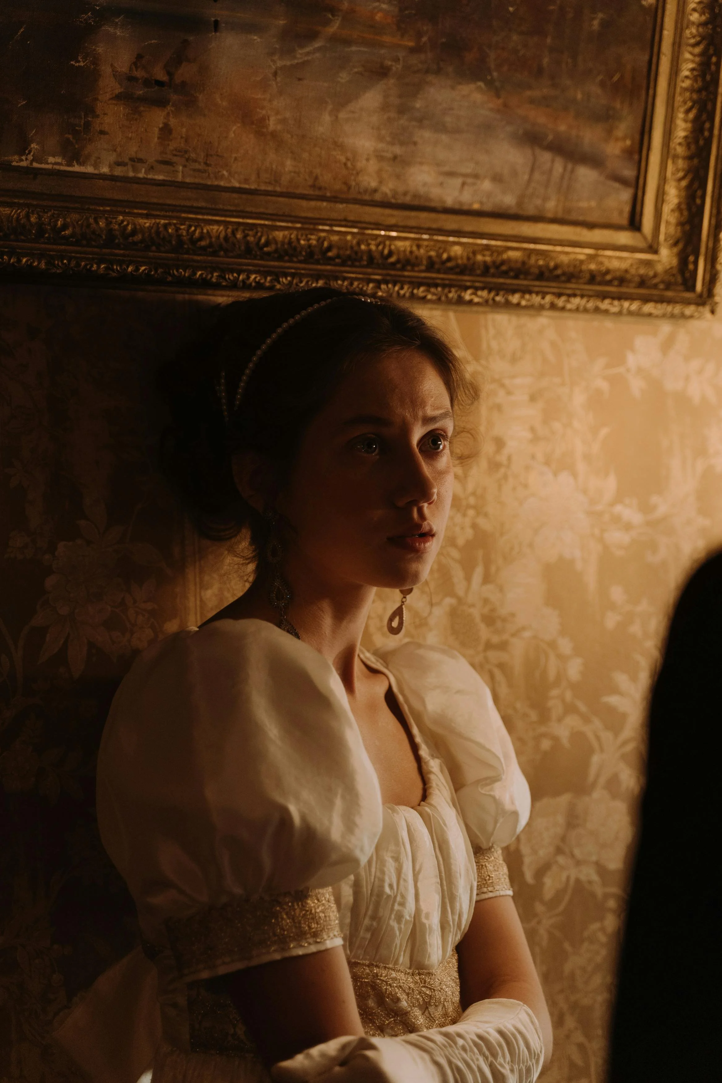 A woman dressed in historical attire with puffed sleeves, ornate jewelry, and gloves, standing against a floral wallpapered wall with a framed painting above her.