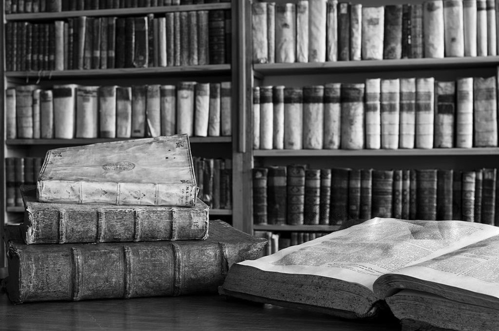 Old books on a table with a large open book in front, in front of a bookshelf filled with more books, black and white photo.