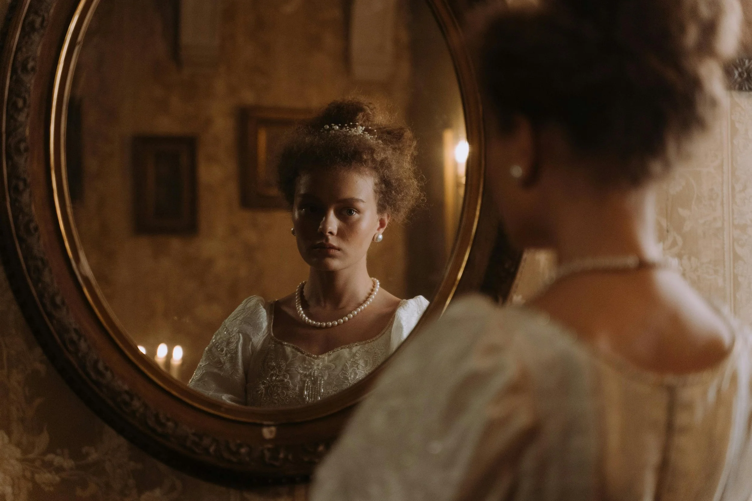 A woman in vintage attire looks at her reflection in an oval mirror. She wears a pearl necklace and earrings, and her hair is styled up with a decorative hairpiece. The background has warm lighting and framed pictures on a patterned wallpapered wall.