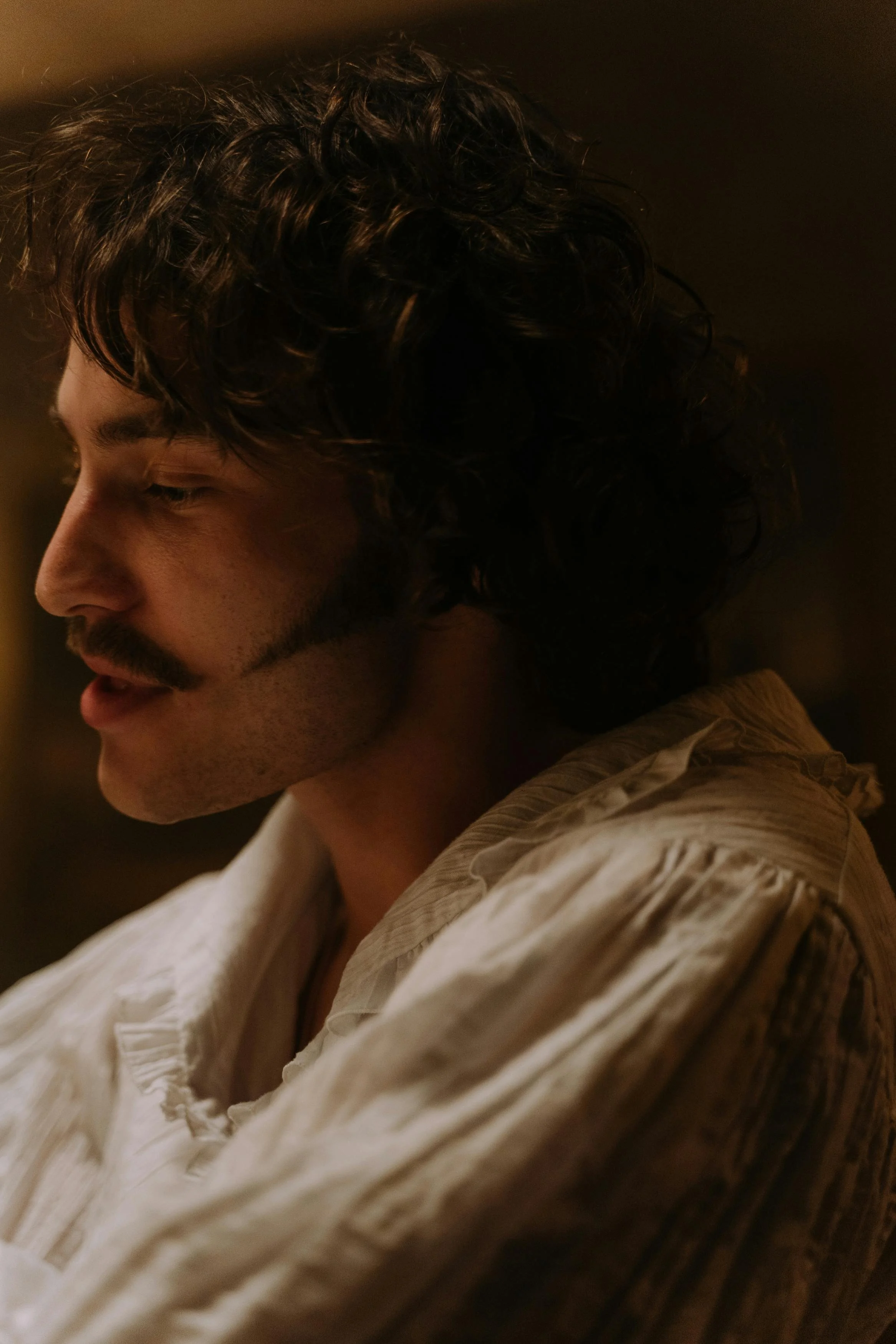 Close-up of a man with curly hair and a mustache, wearing a cream-colored shirt, looking down in a dimly lit environment.