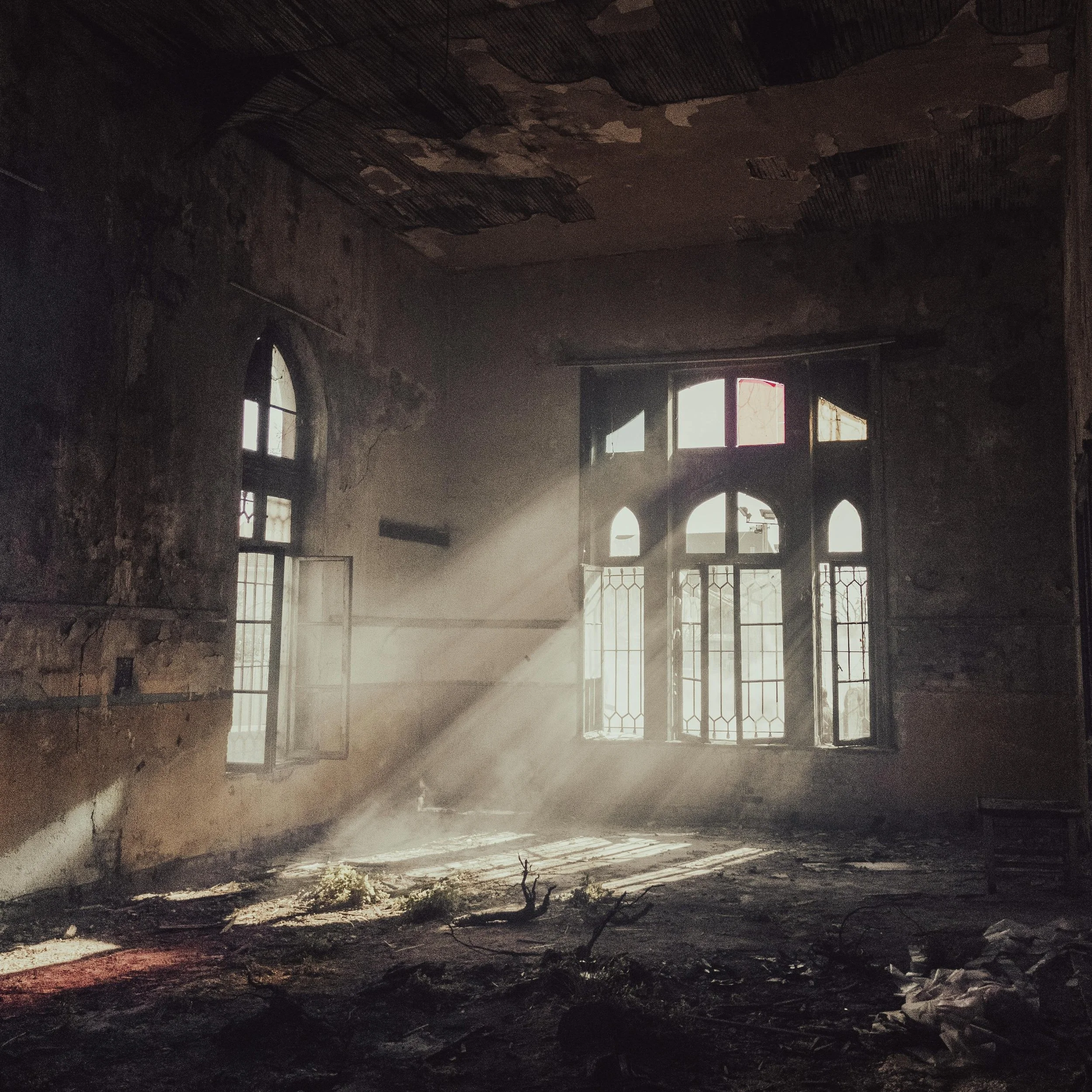 Sunlight streams through the broken windows of a dilapidated, abandoned building with peeling paint and debris on the floor.