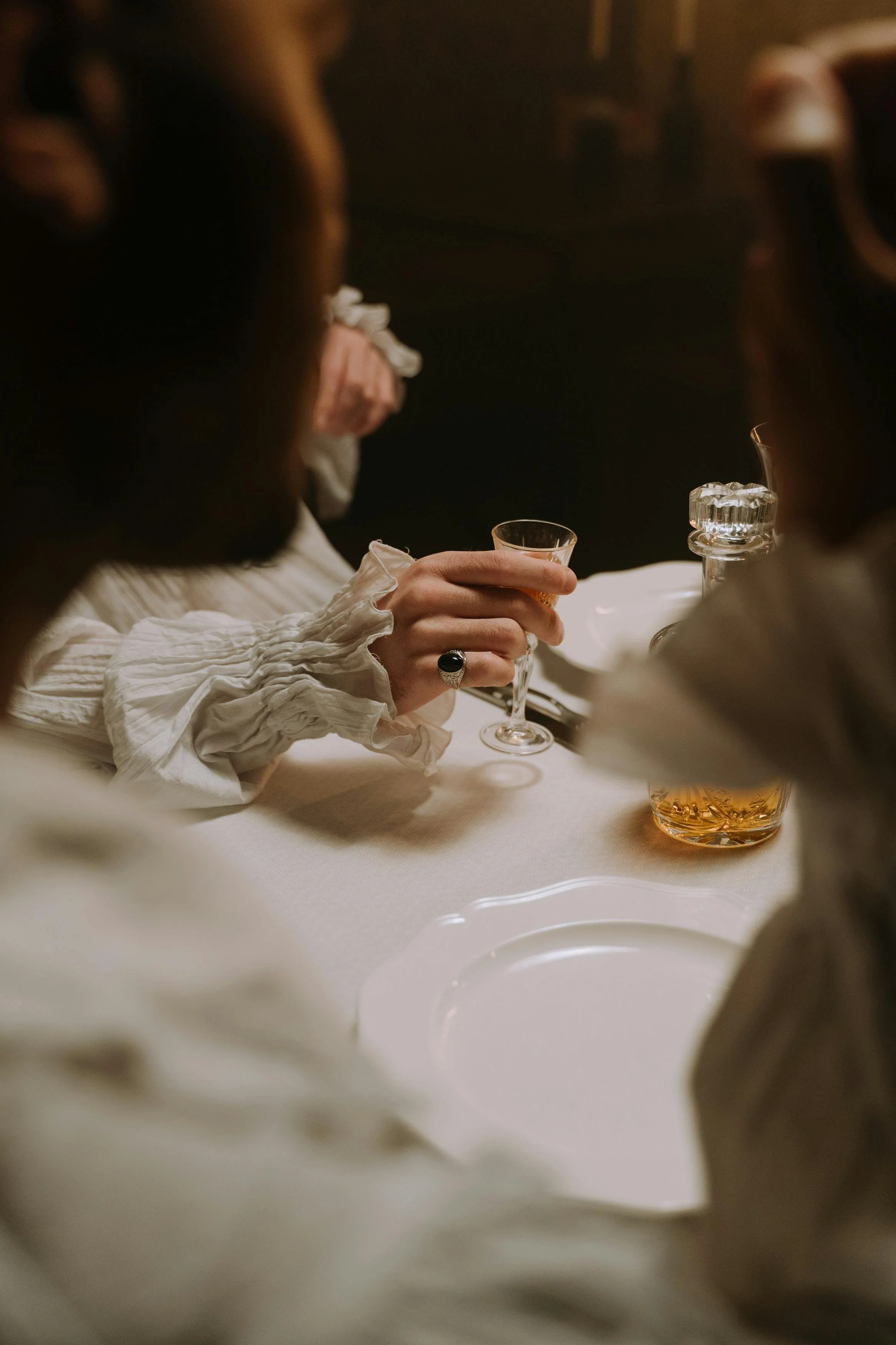 People sitting around a table, with one person holding a glass of liquor, dressed in vintage clothing.