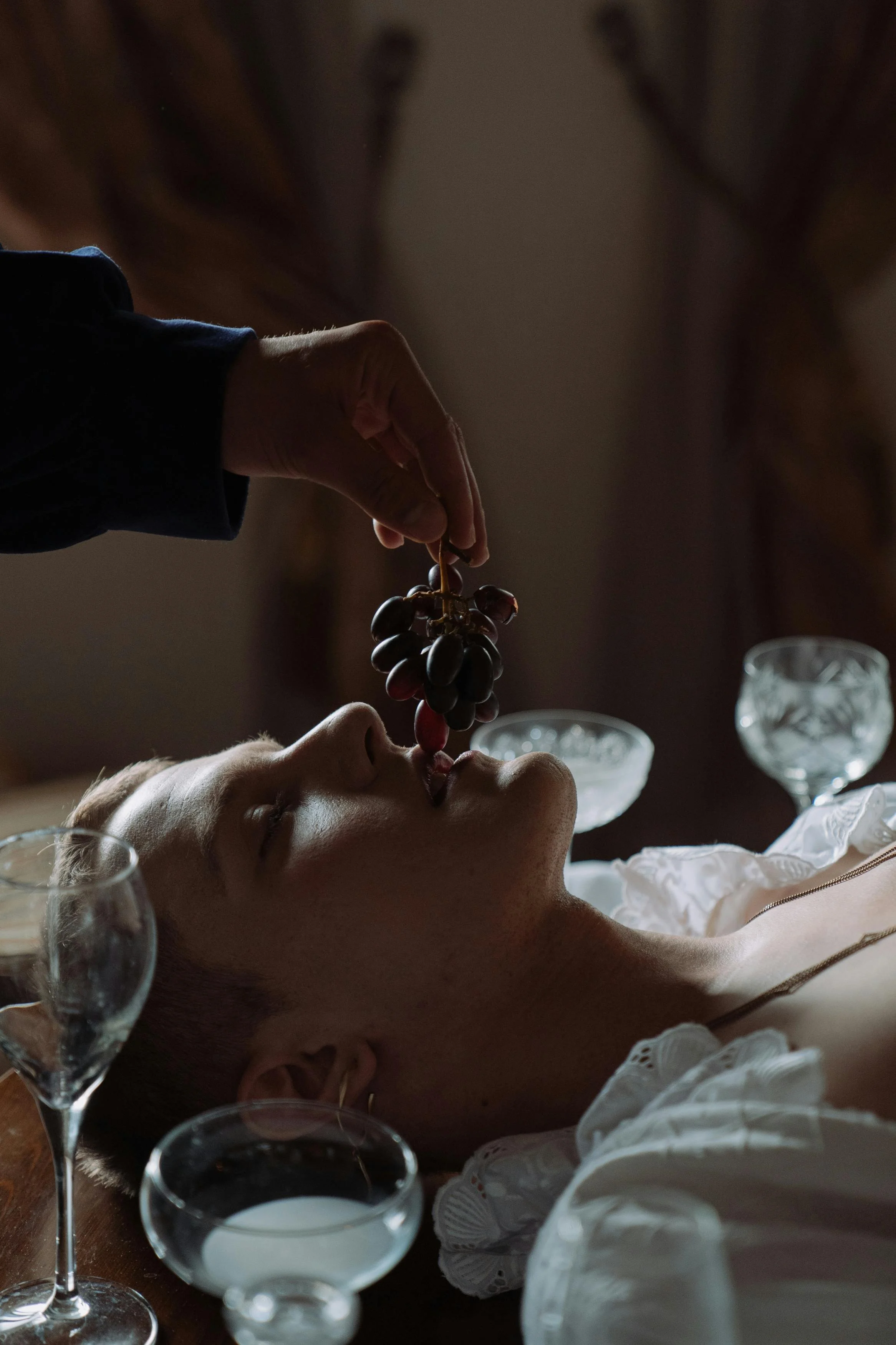 A person lying on a table with eyes closed, being fed grapes by another person, with glassware on the table.