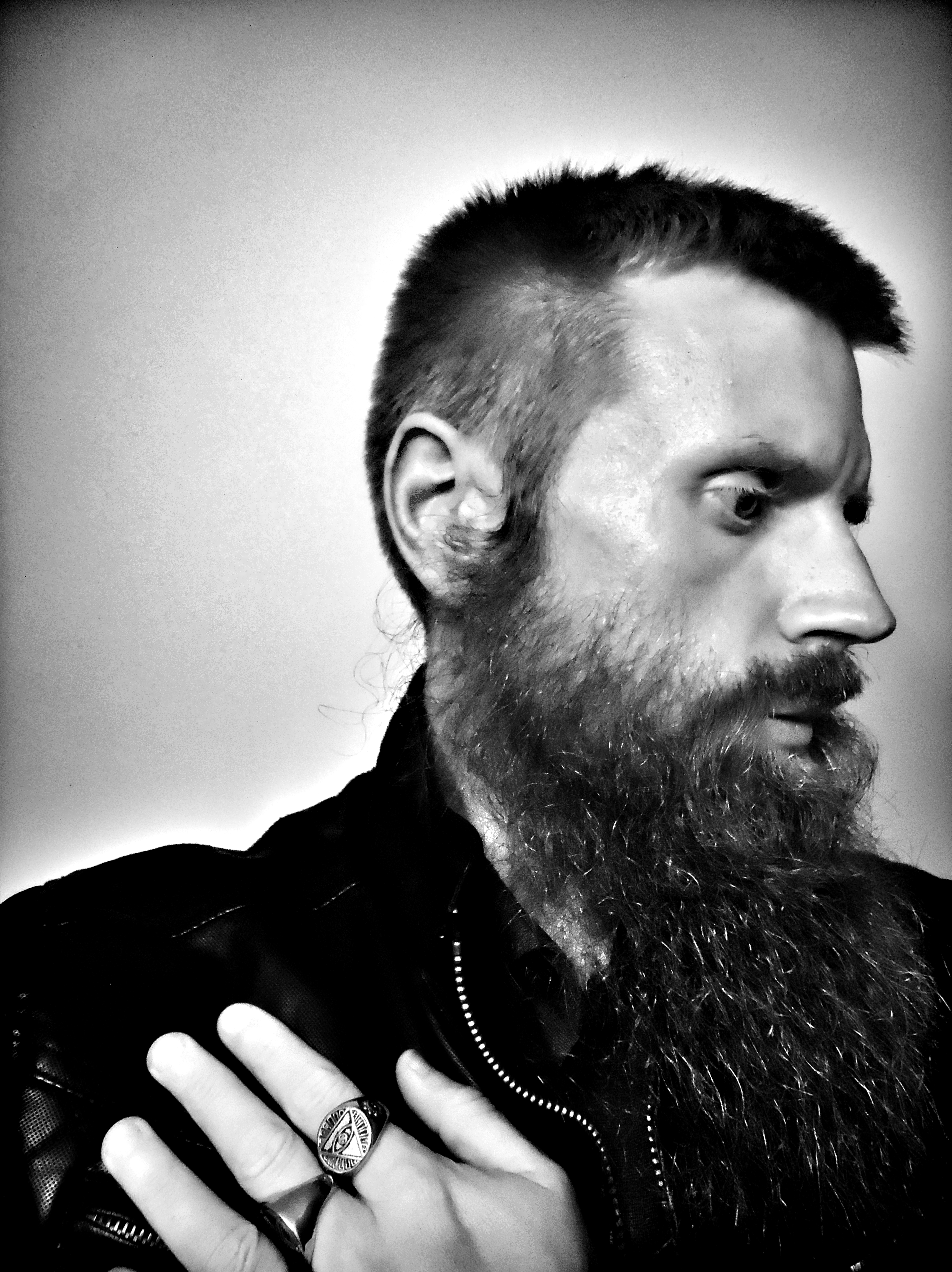 Black and white portrait of a man with a beard and short hair, wearing a leather jacket, and displaying rings on his fingers, one with an eye symbol.