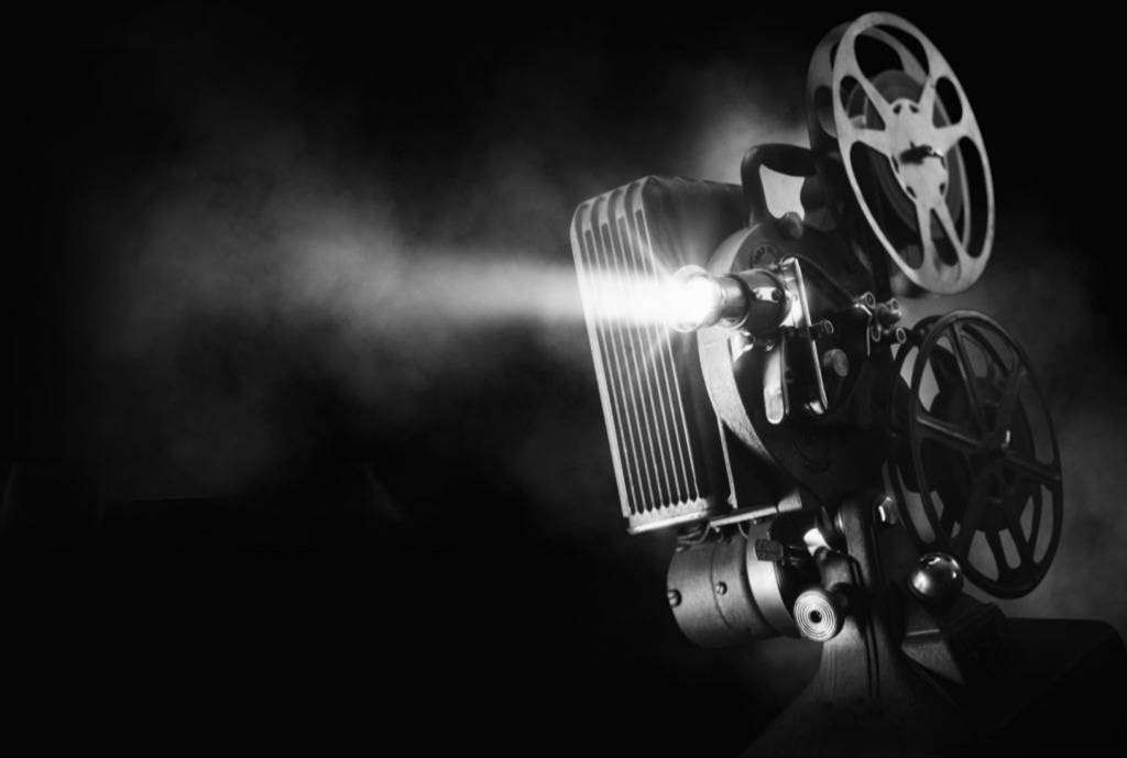 A vintage film projector emitting light with reel wheels, casting a beam of light into darkness.