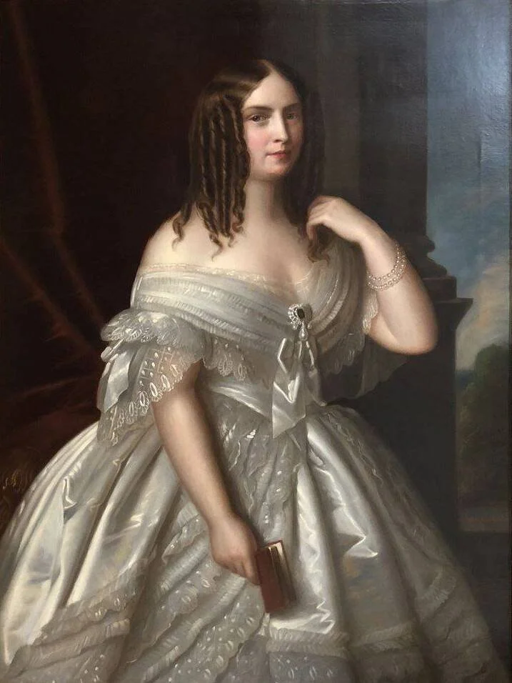 A young woman with brown hair in an elegant, historic dress standing indoors, holding a small purse in her right hand, wearing a pearl bracelet, with a neutral expression.
