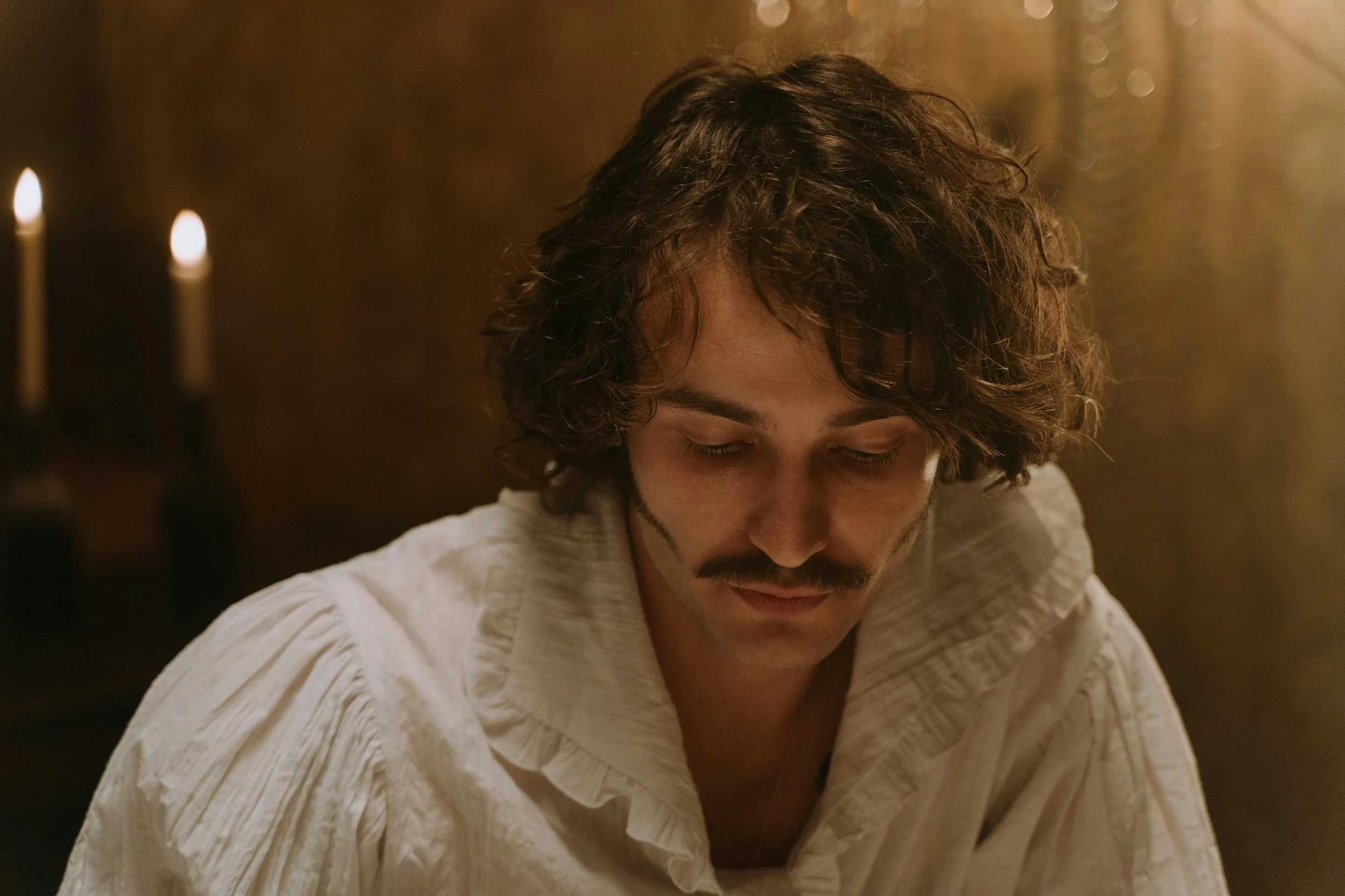 A man with curly hair and a mustache looks down, wearing a white shirt, with a warm, dimly lit background and candles.