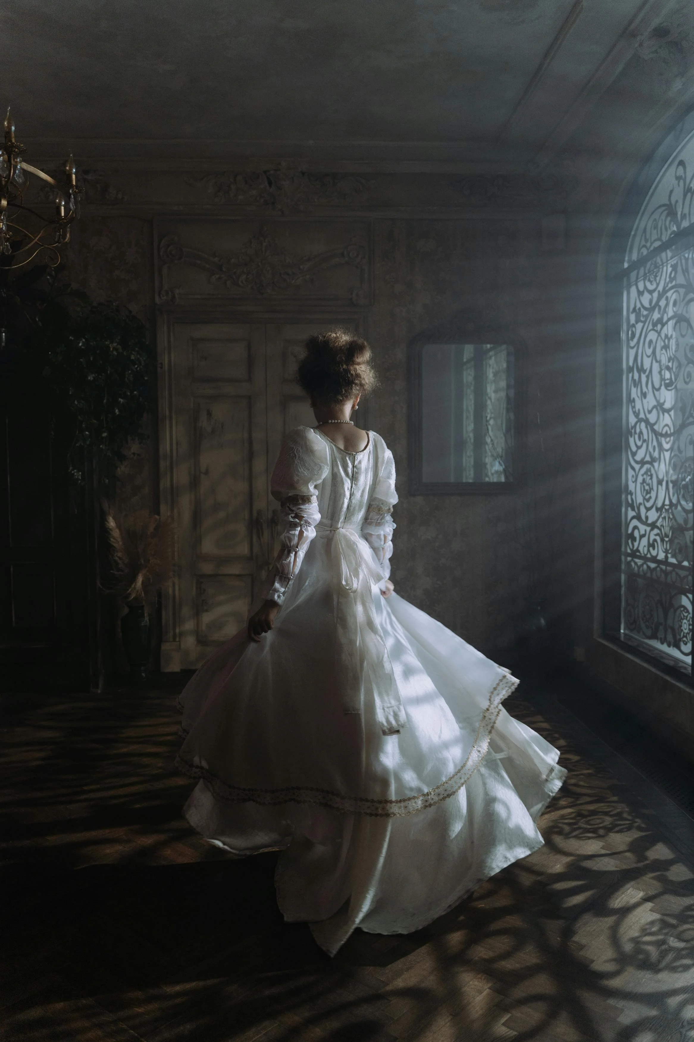 A woman in a vintage white dress standing in a dimly lit room with sunlight streaming through large, ornate window panels, casting decorative shadows on the wooden floor.