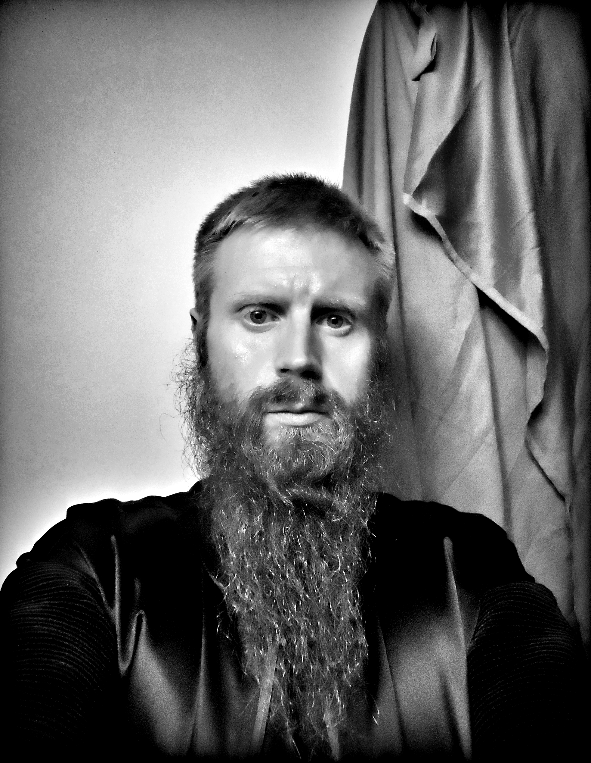 Black-and-white self-portrait of Jono Borden facing the viewer head-on, serious expression, long beard, black satin jacket, drapery behind him, taken of himself in 2020.
