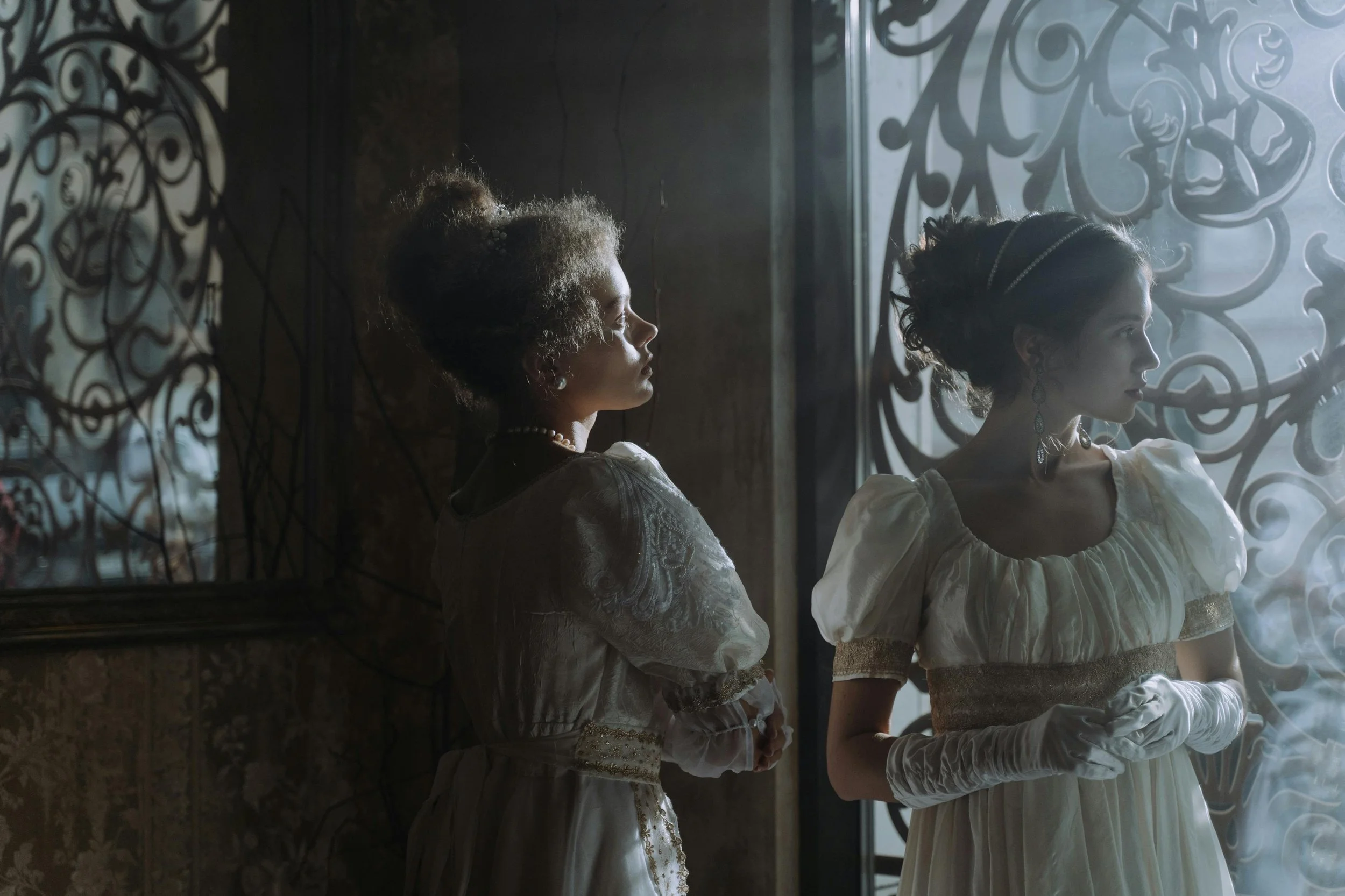 Two women dressed in vintage white dresses and gloves stand near a window with decorative wrought ironwork, with one woman facing left and the other facing right, both illuminated by soft, dramatic lighting.