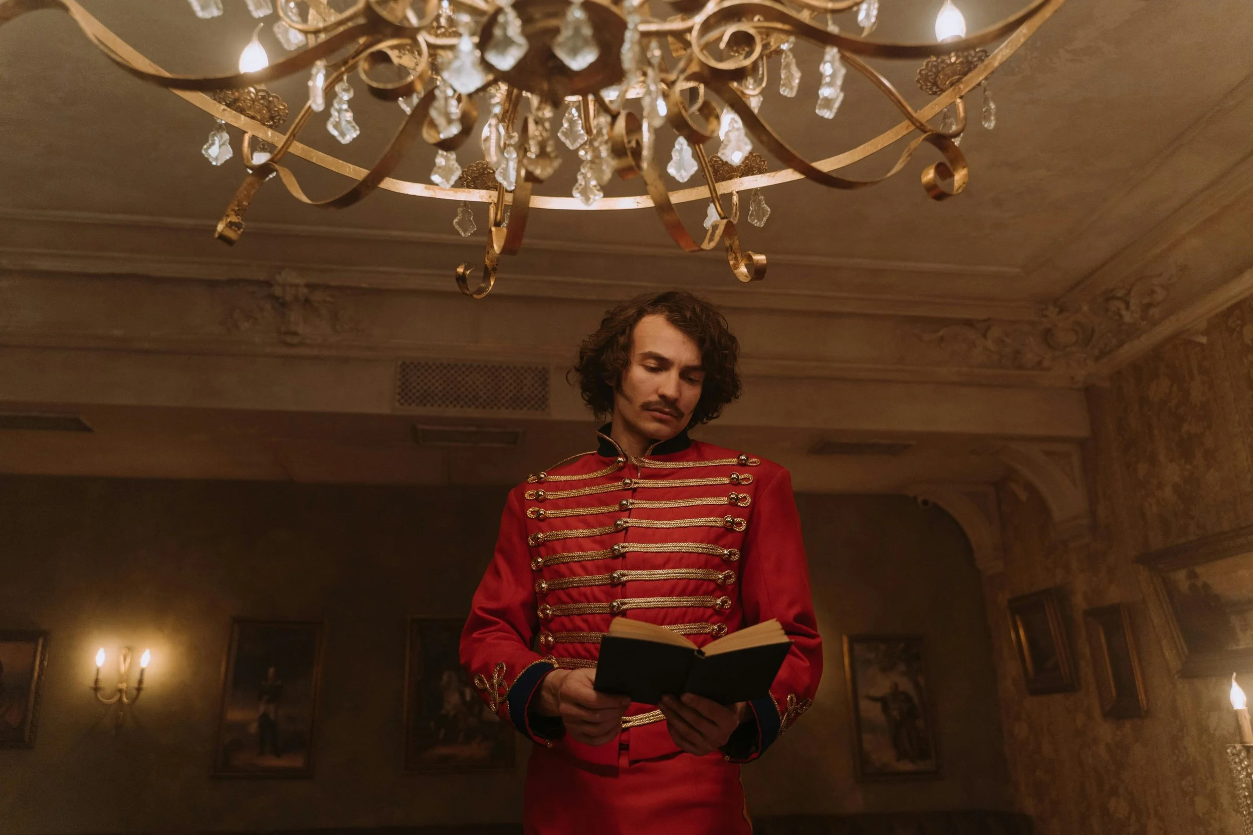 A man dressed in a red military-style jacket with gold embellishments stands inside a dimly lit room, reading a small black book. The room has ornate wallpaper, framed pictures on the walls, and a chandelier hanging from the ceiling.