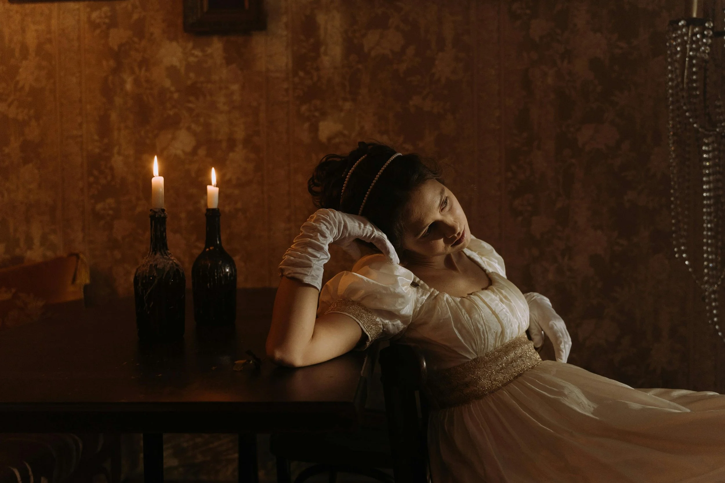 A woman in a vintage dress reclines at a dark wooden table with two lit candles in black glass holders; she is resting her head on her hand, gazing thoughtfully, in a dimly lit, ornate room with patterned wallpaper.