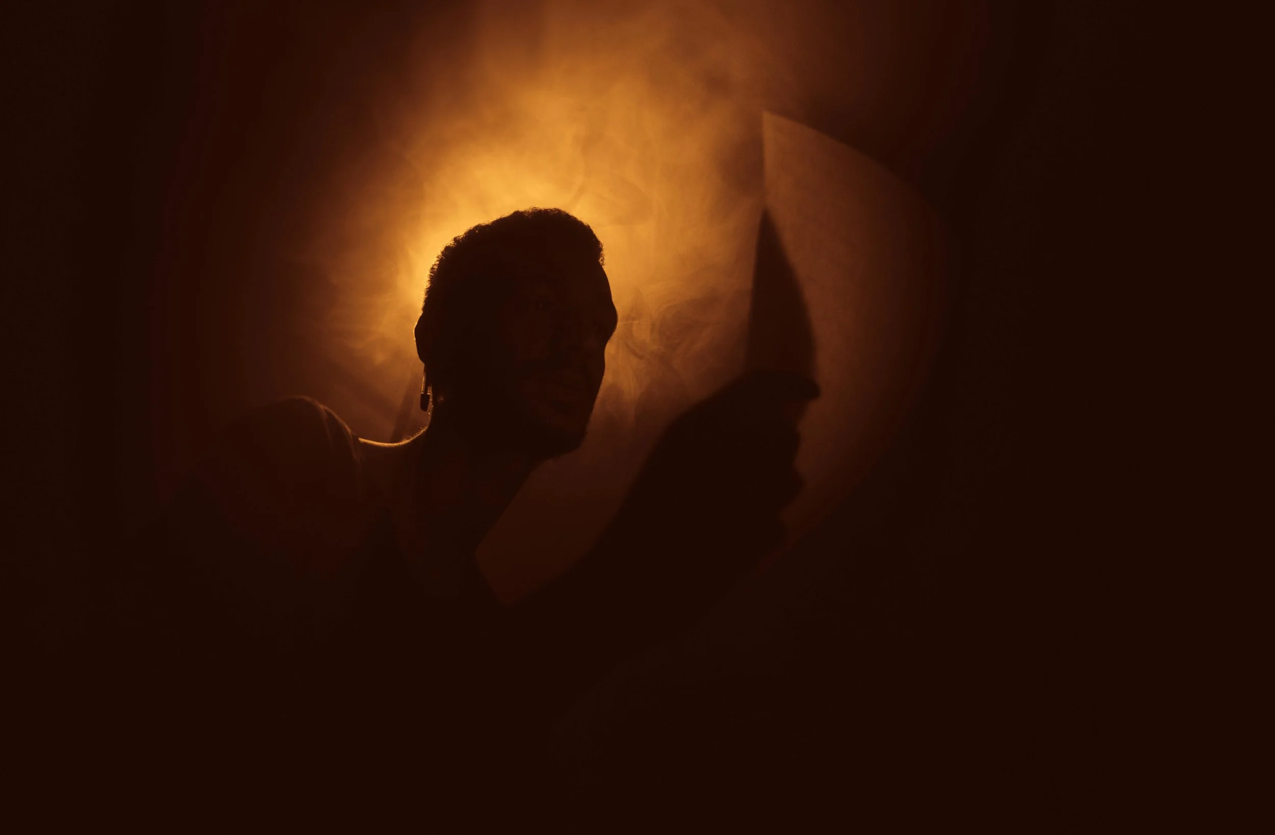 Silhouette of a person with short hair and earrings, backlit by warm, smoky light, creating a shadowed profile and surrounding atmospheric effects.
