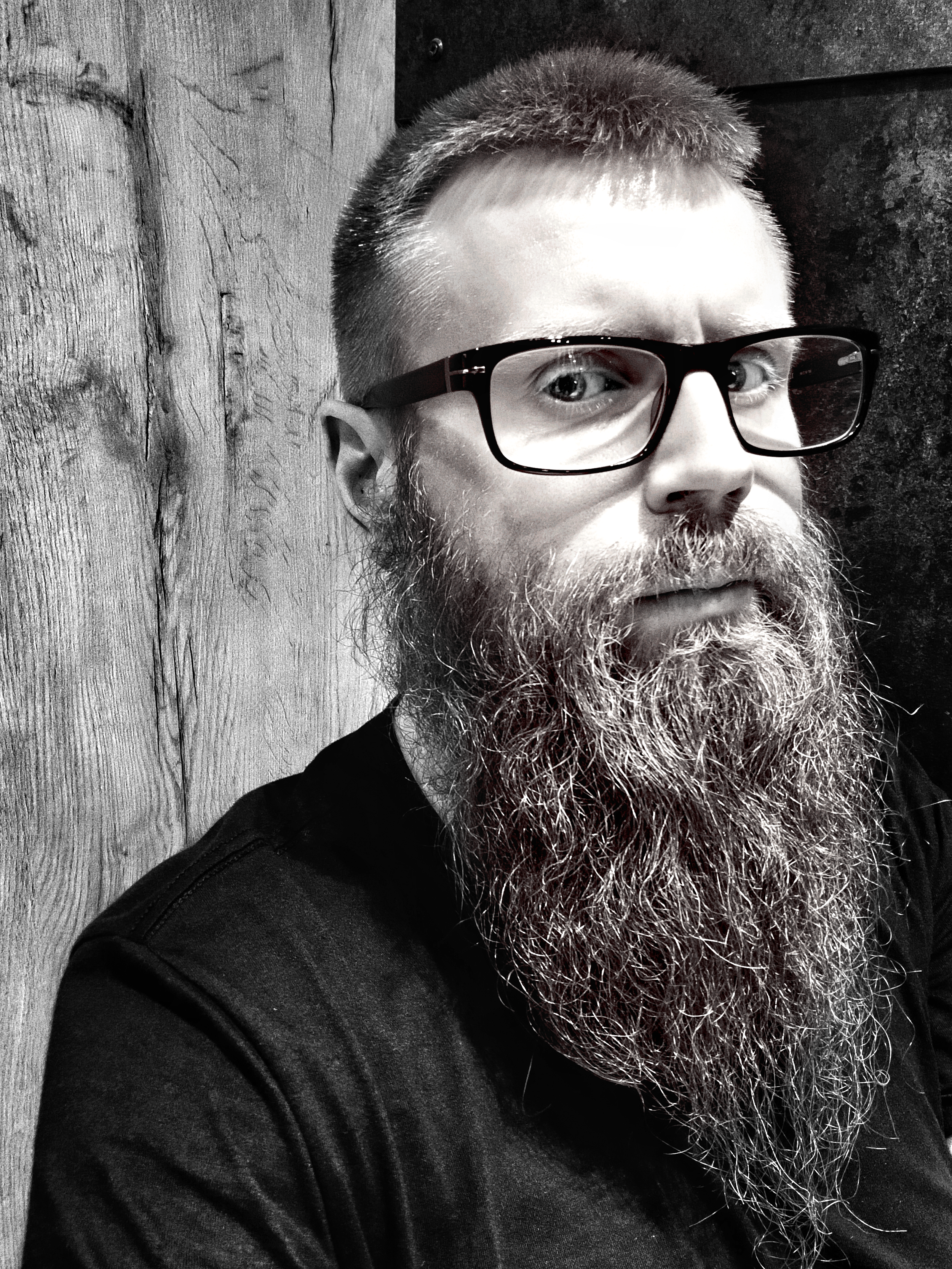 Close-up black and white photo of a man with glasses and a long beard, sitting against a wooden wall.