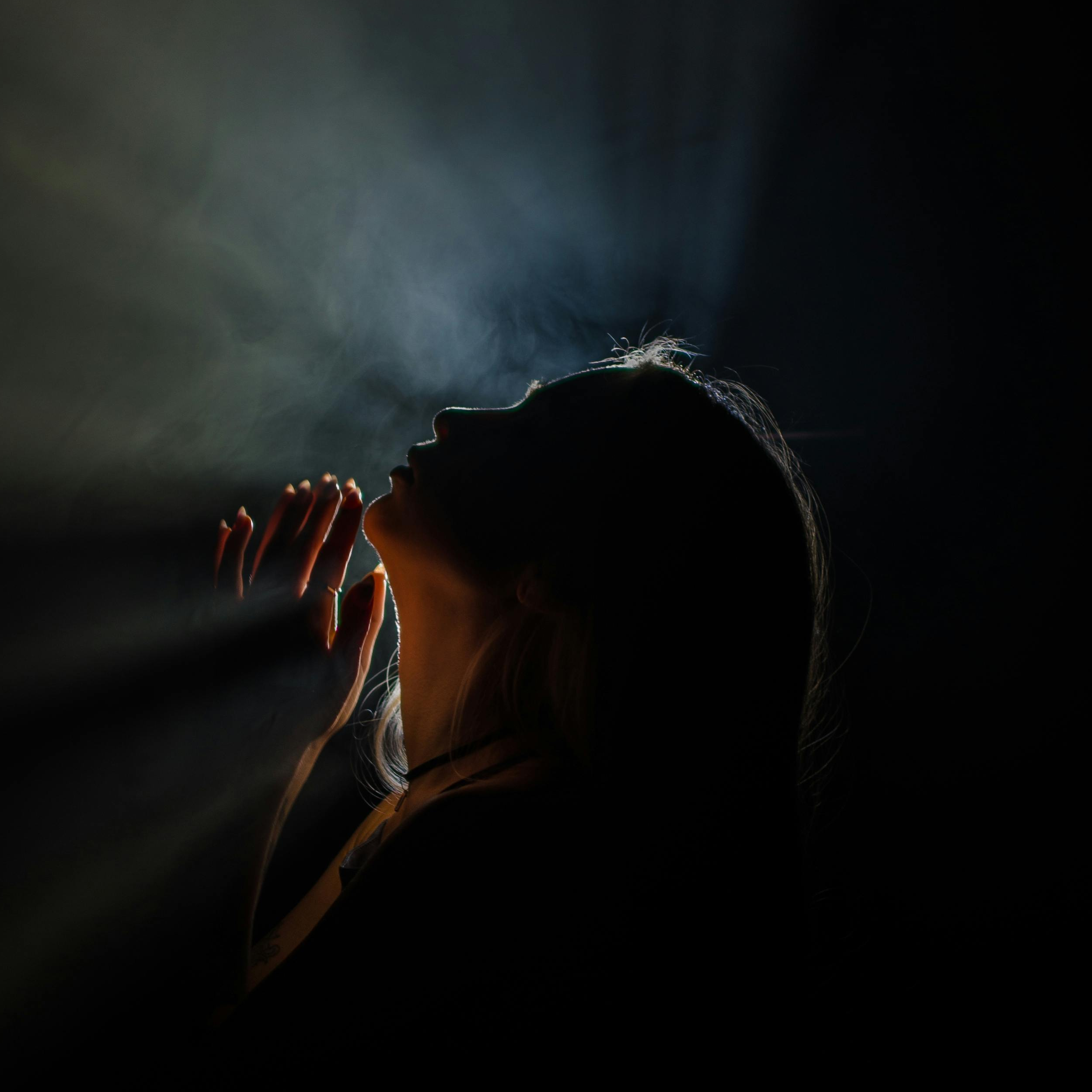 Silhouette of a woman with her eyes closed and hands near her face, backlit with a subtle glow, against a dark background.