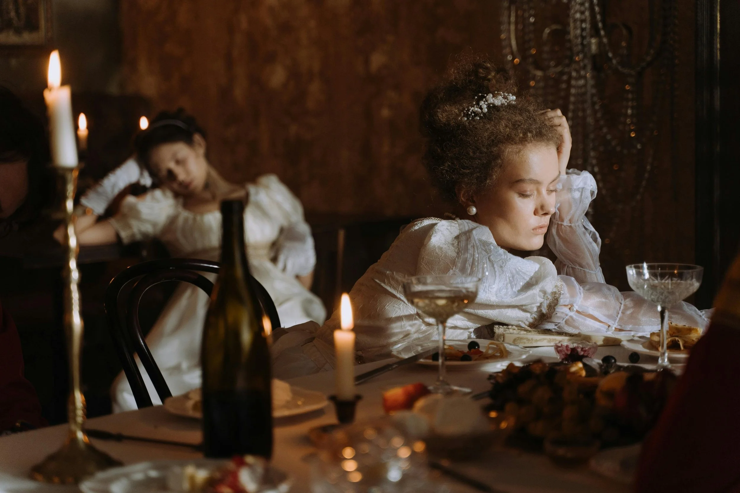 Two young girls in vintage-style white dresses sitting at a decorated dinner table with lit candles, with one girl resting her head on her hand and appearing bored or tired.