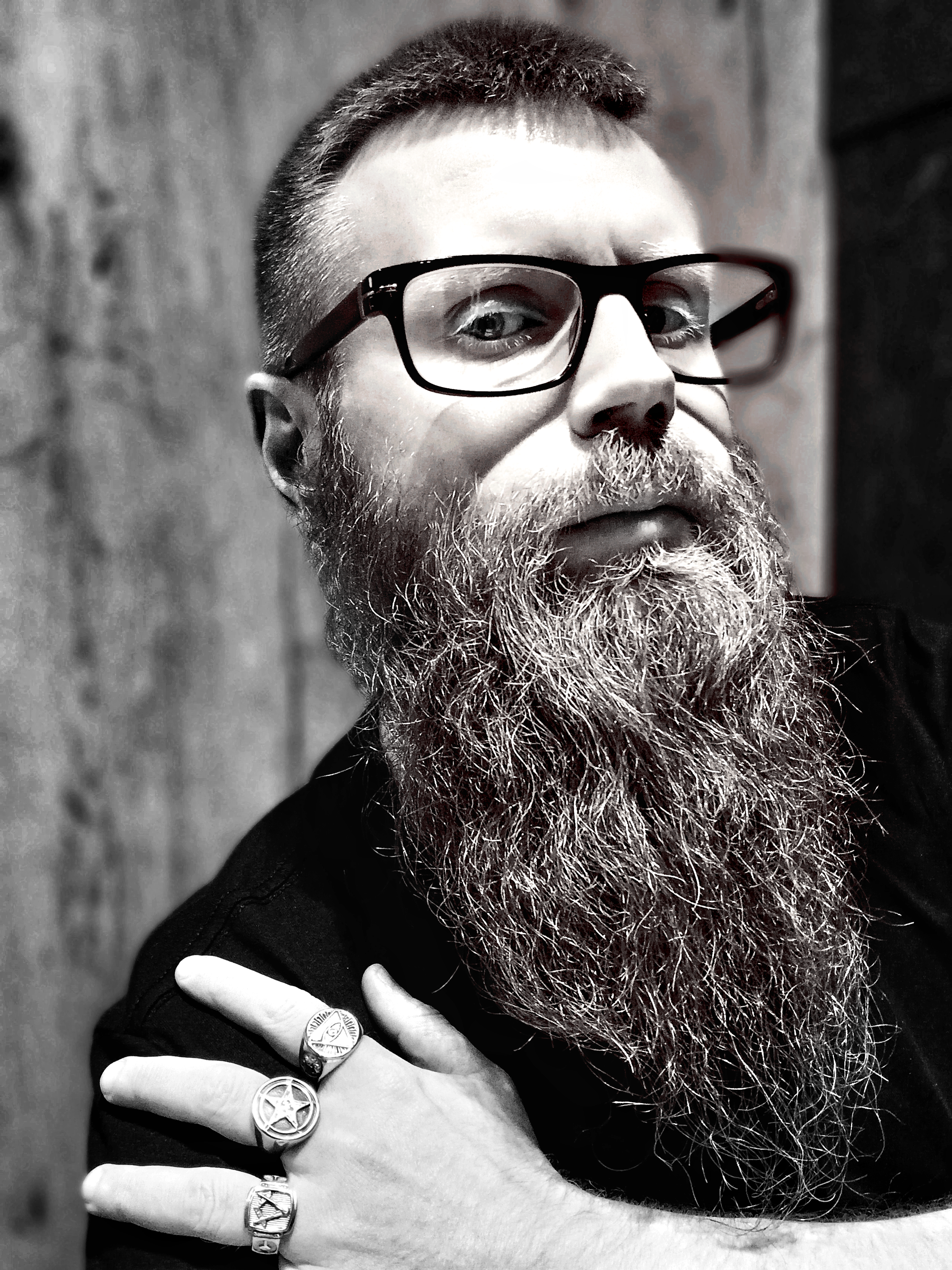A man with glasses and a long beard, wearing rings on his fingers, looking at the camera with a serious expression.