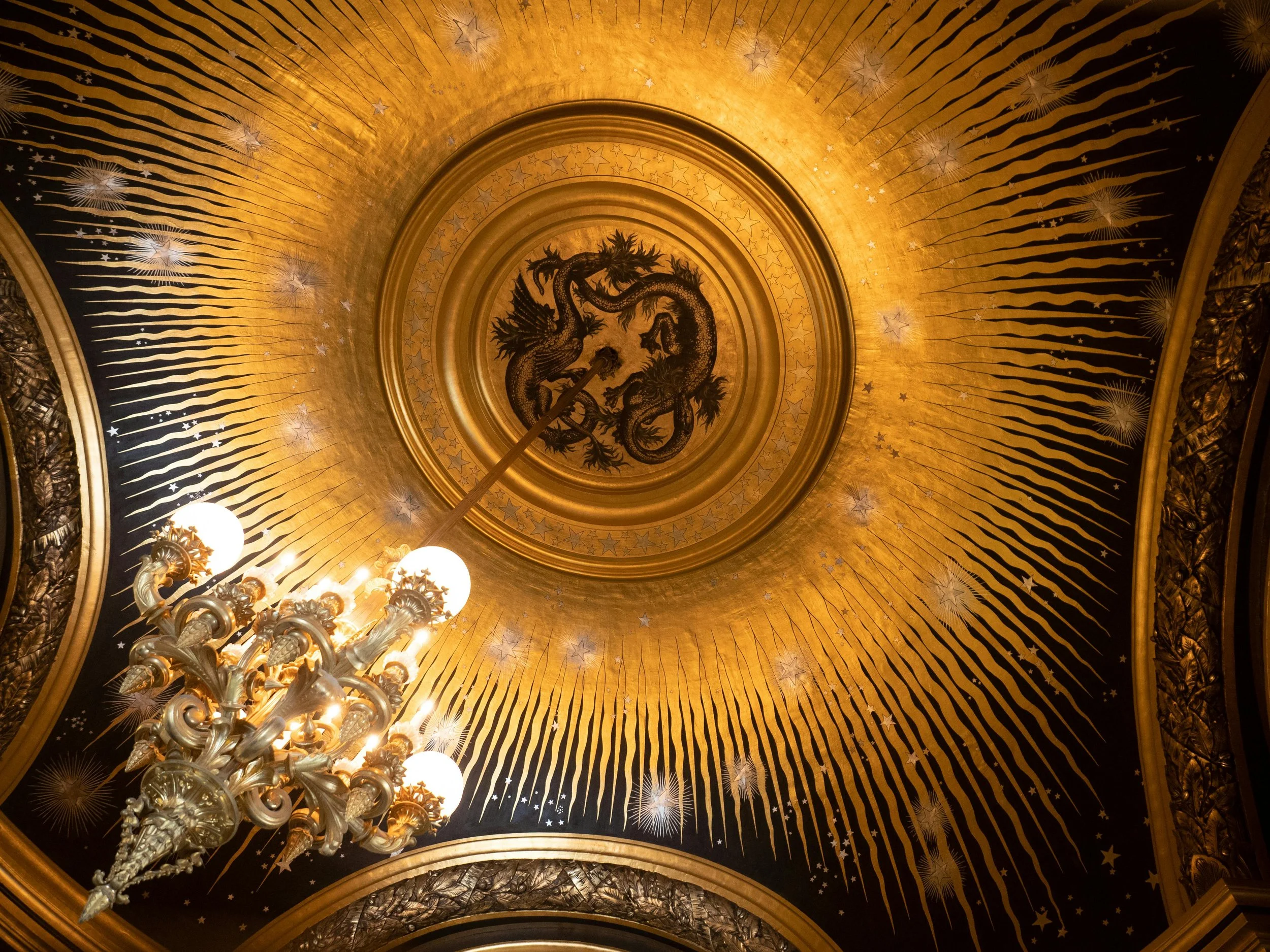 The ornate ceiling is decorated with gold and black patterns, featuring a central emblem of two dragons intertwined, surrounded by radiant rays and star motifs, with a chandelier hanging below.