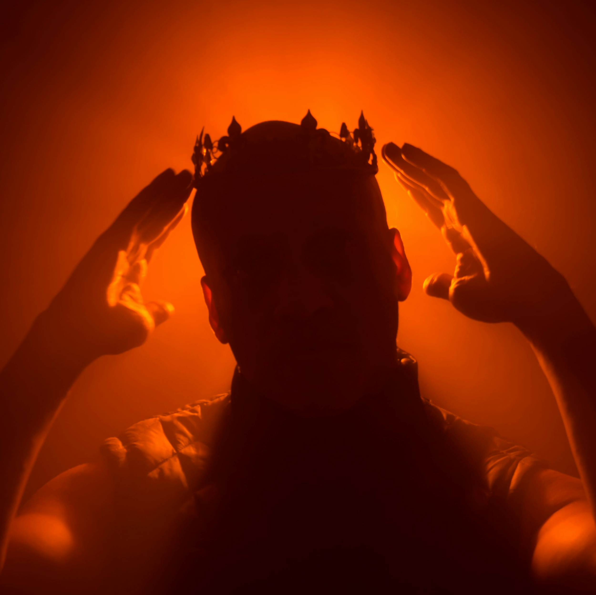 Silhouette of a man adjusting a crown on his head against a warm orange background.