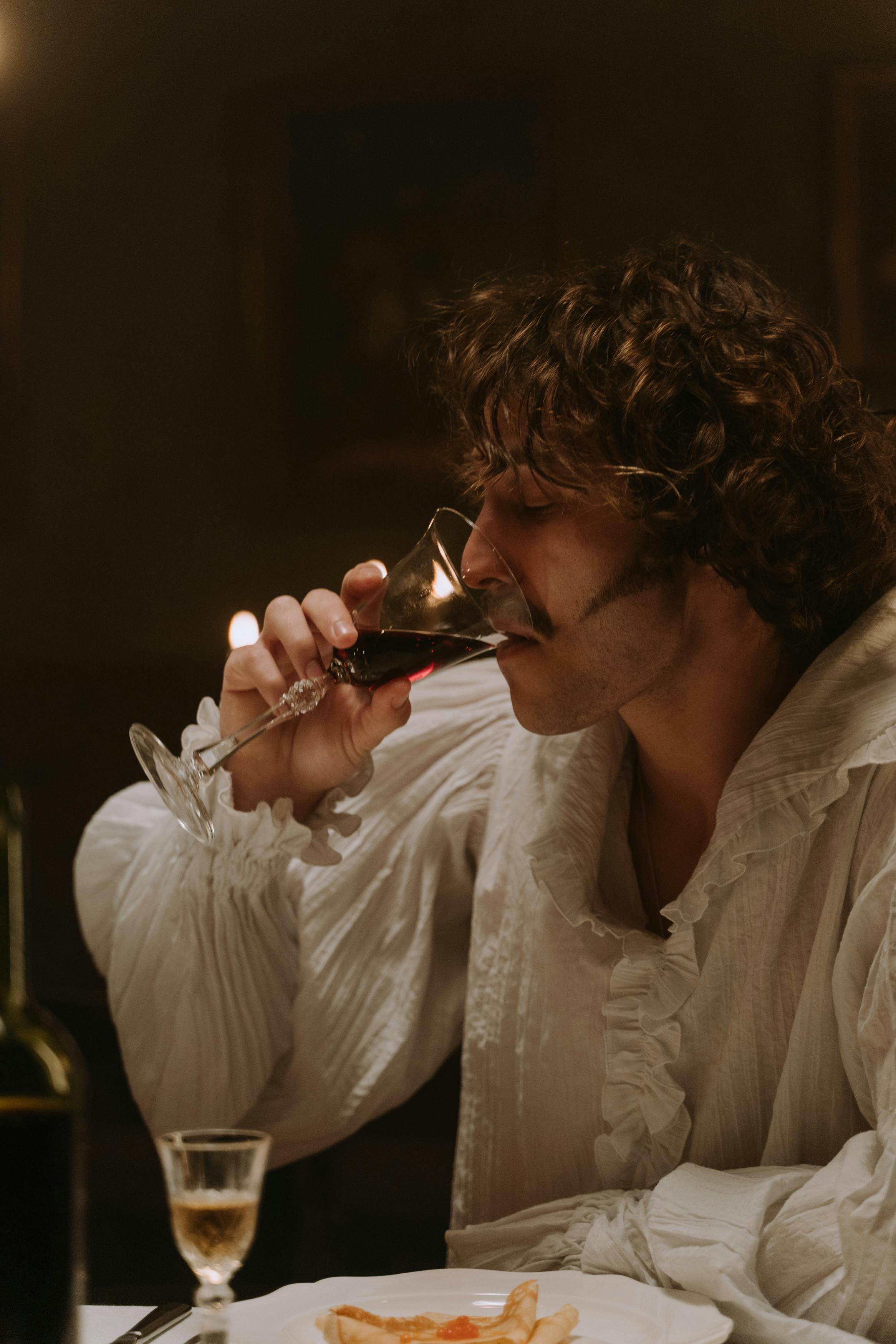 A man with curly hair and a mustache is drinking red wine from a glass in a dimly lit setting.