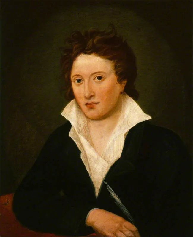 Portrait of a young man with curly brown hair, wearing a white shirt with a high collar and a black jacket, seated against a dark background.