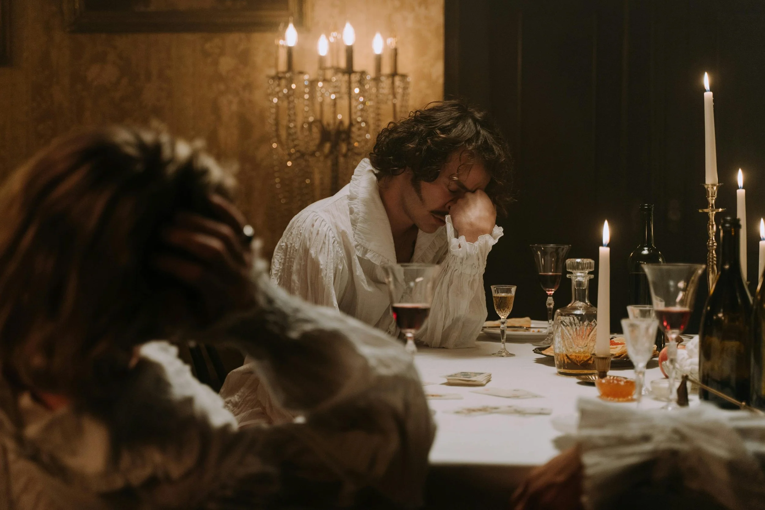 A person with curly hair wearing a white shirt sitting at a table with a hand on their forehead, looking distressed, with lit candles and glassware on the table, in a dimly lit room.