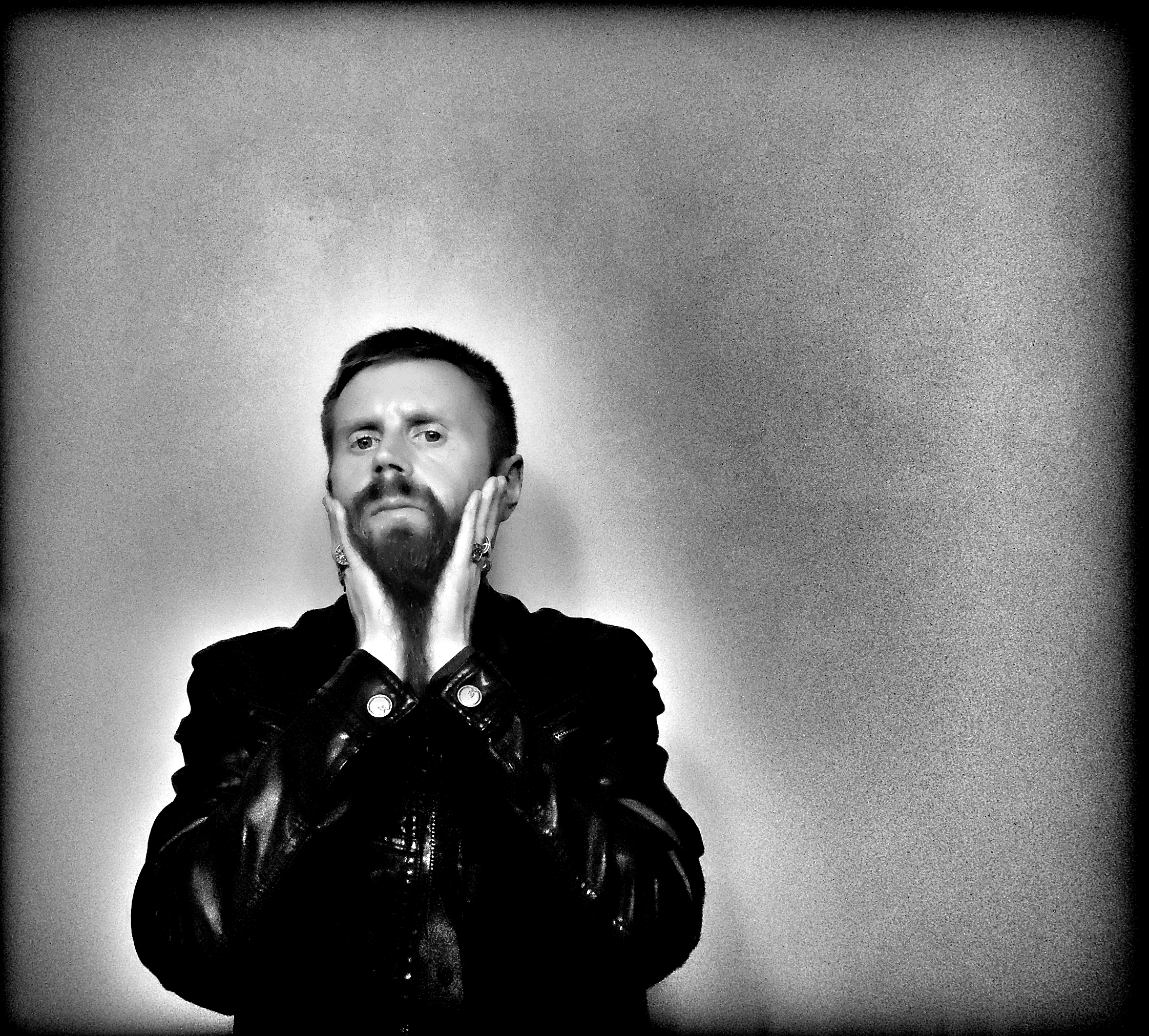 Black-and-white self-portrait of Jono Borden wearing a black leather jacket, wearing many large silver rings, holding his face with palms outstretched, pulling down his long bear, facing the viewer head-on, taken of himself in 2020.