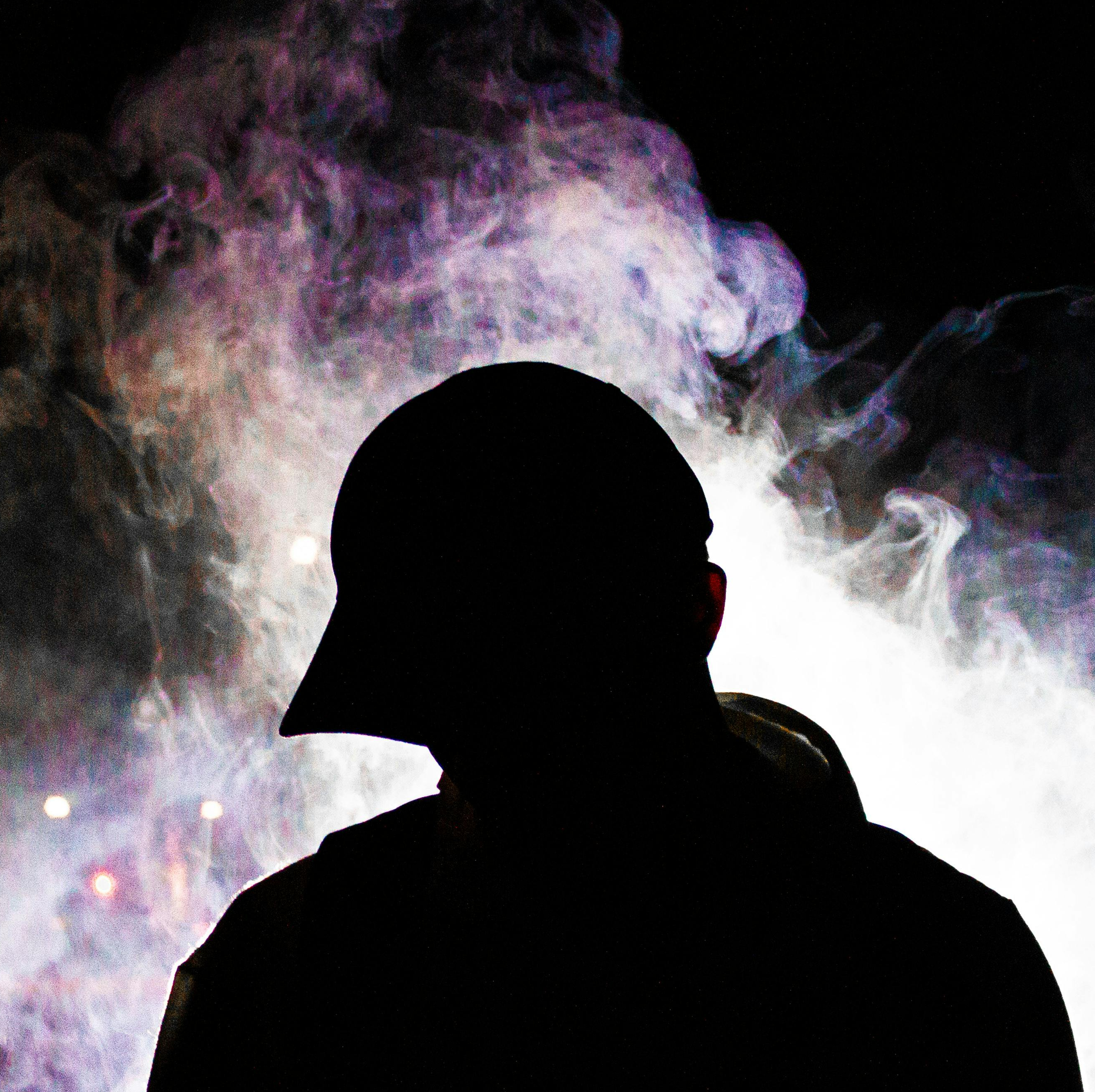Silhouette of a person wearing a cap in front of colorful smoke or vapor at night.