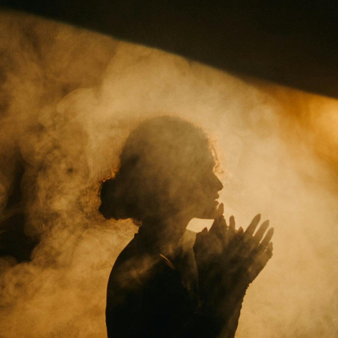 Silhouette of a person with hands clasped together, praying, surrounded by a foggy, dimly lit atmosphere.
