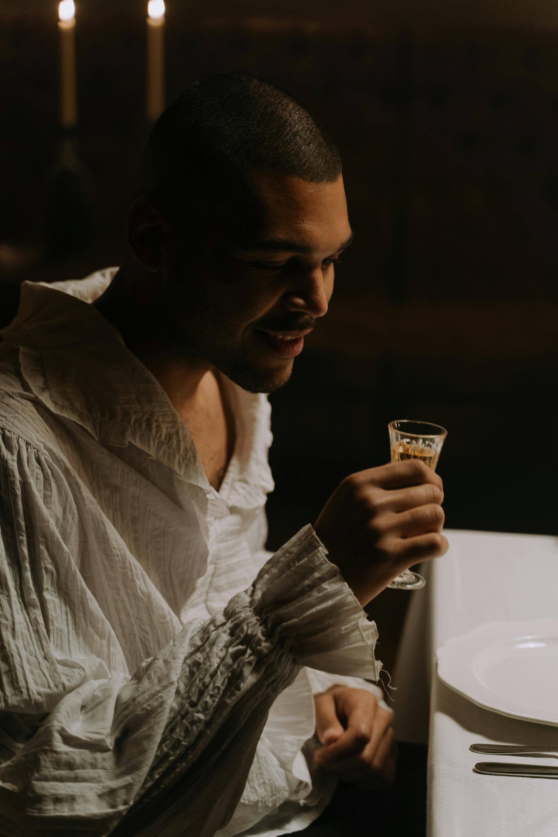 A man in a white shirt sitting at a dining table, holding a glass of what appears to be a drink, with candles in the background.