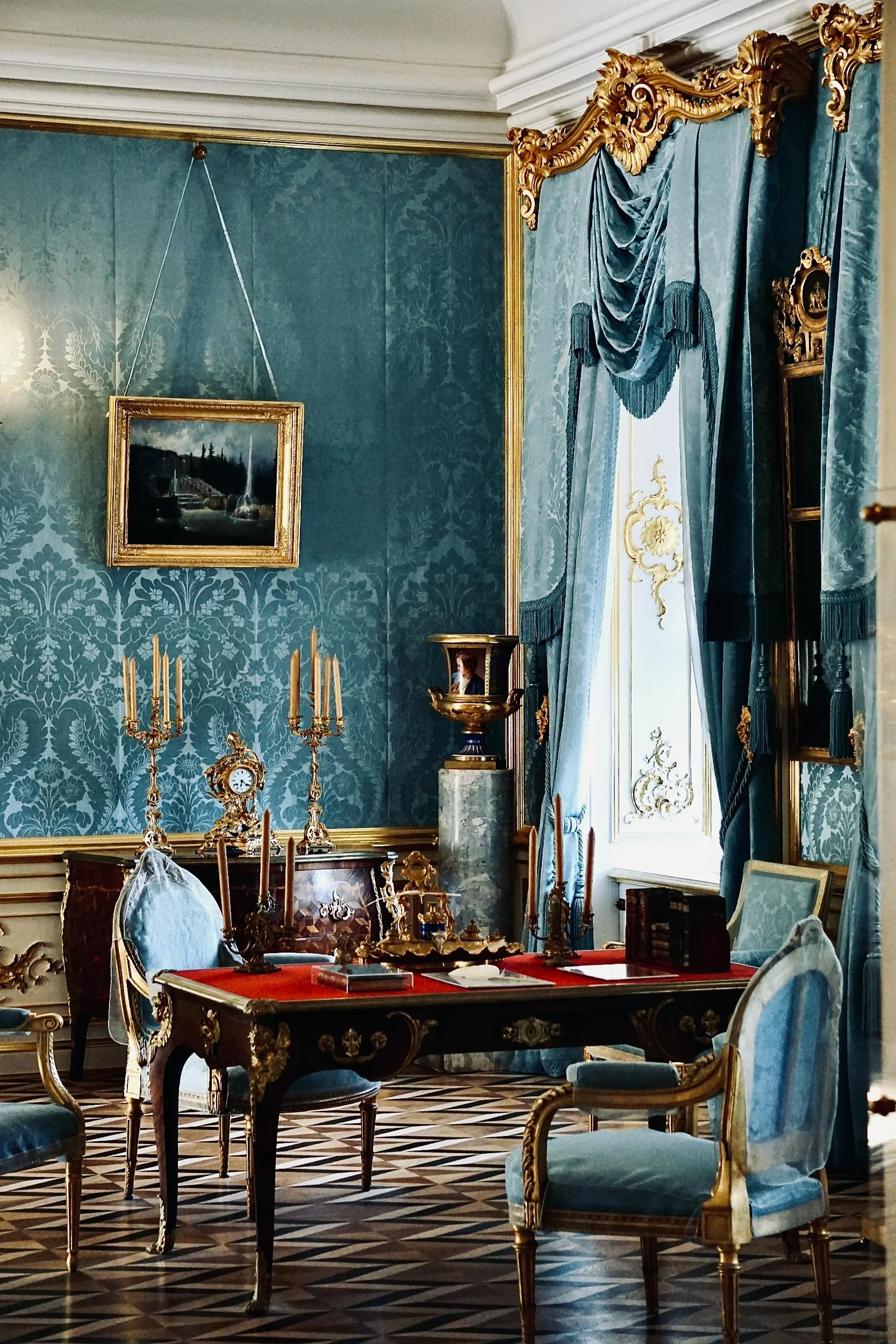 Room with blue wallpaper, gold accents, ornate curtains, a desk with papers, chairs, candelabras, a painting, and decorative items in a luxurious, historic style.
