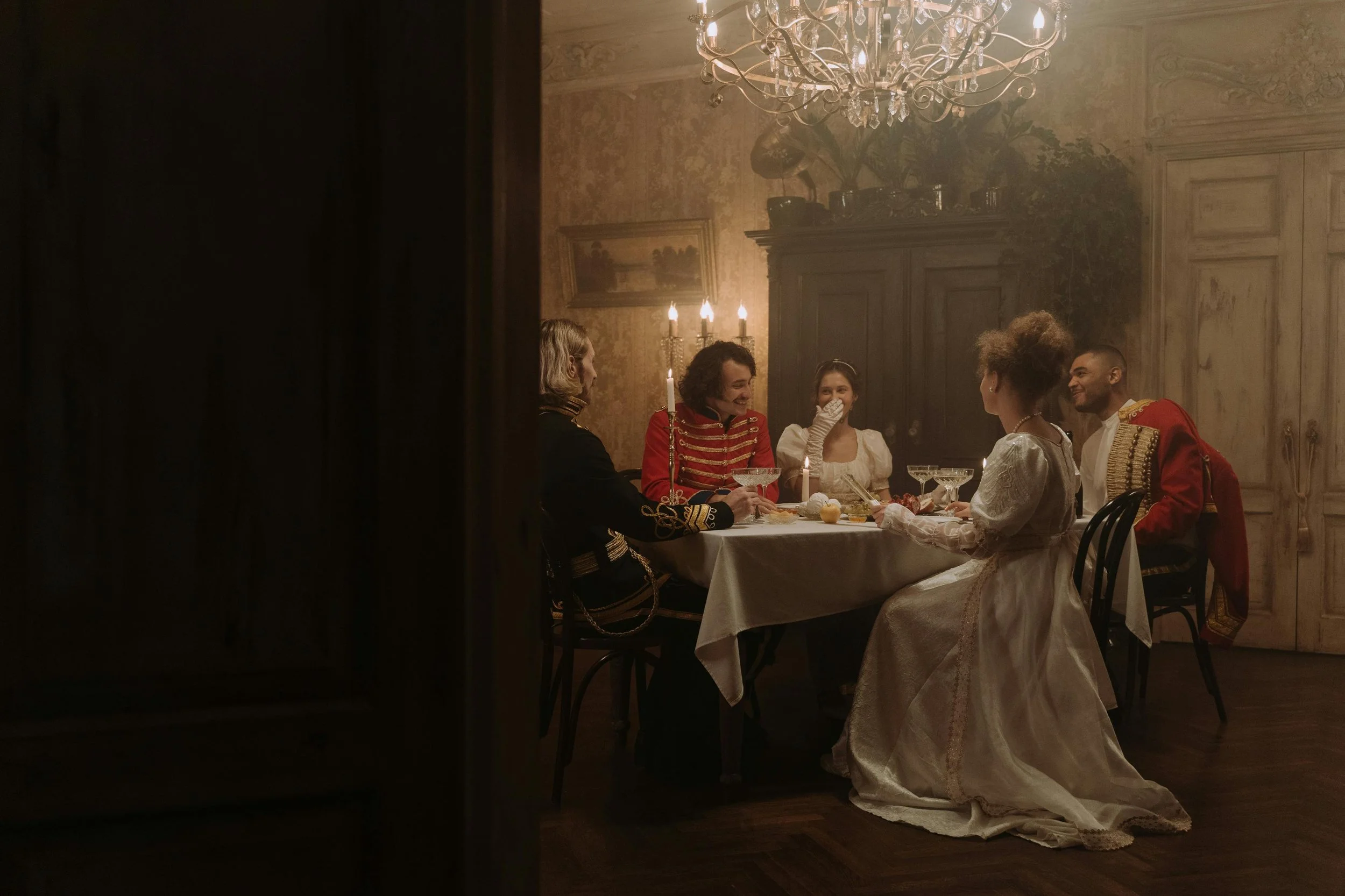 People dressed in historic costumes enjoying a dinner party in a vintage, dimly lit dining room with chandelier and candles.