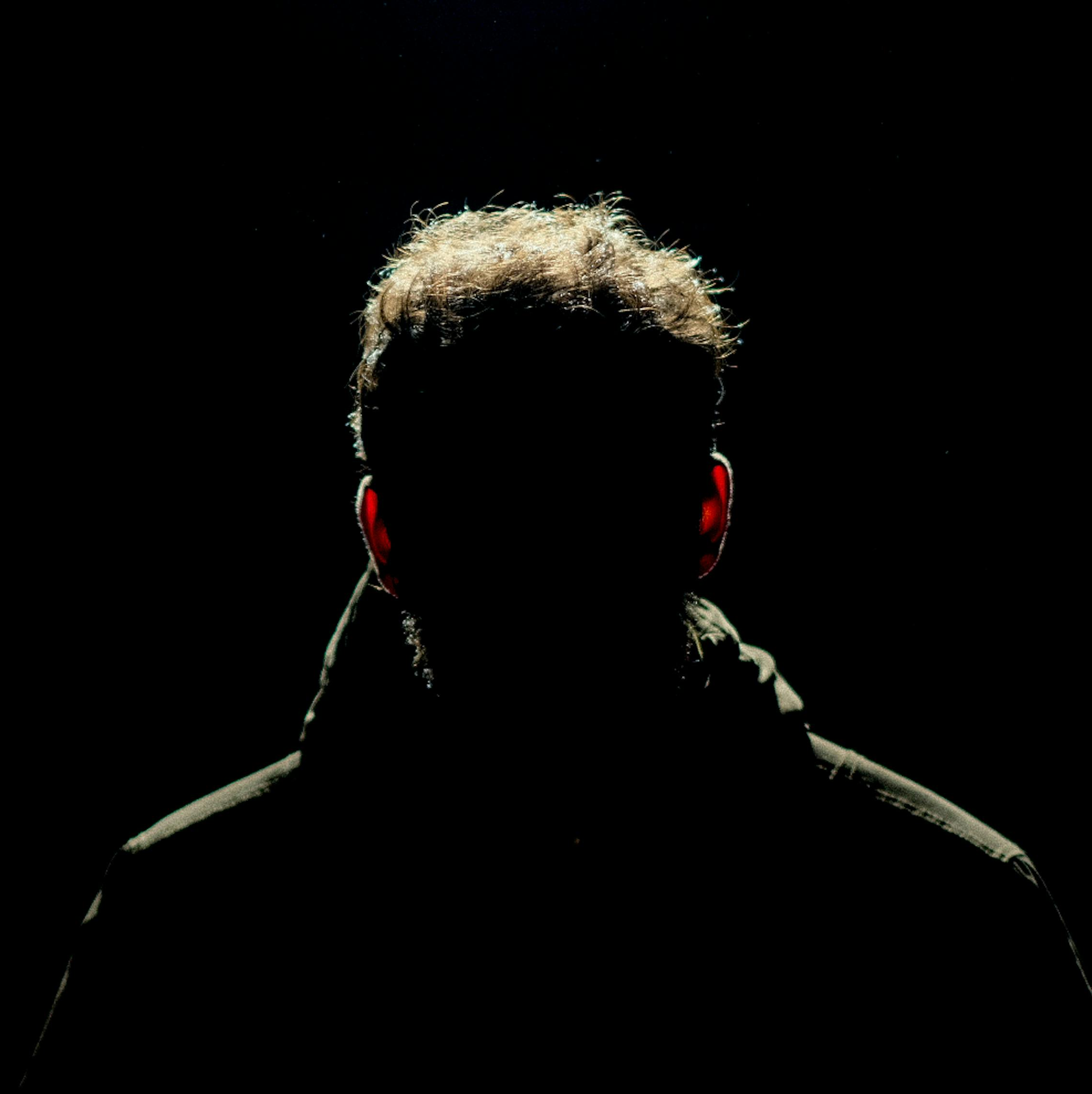 Silhouette of a man with short, curly hair, wearing red headphones, against a dark background, with backlighting highlighting his hair and shoulders.