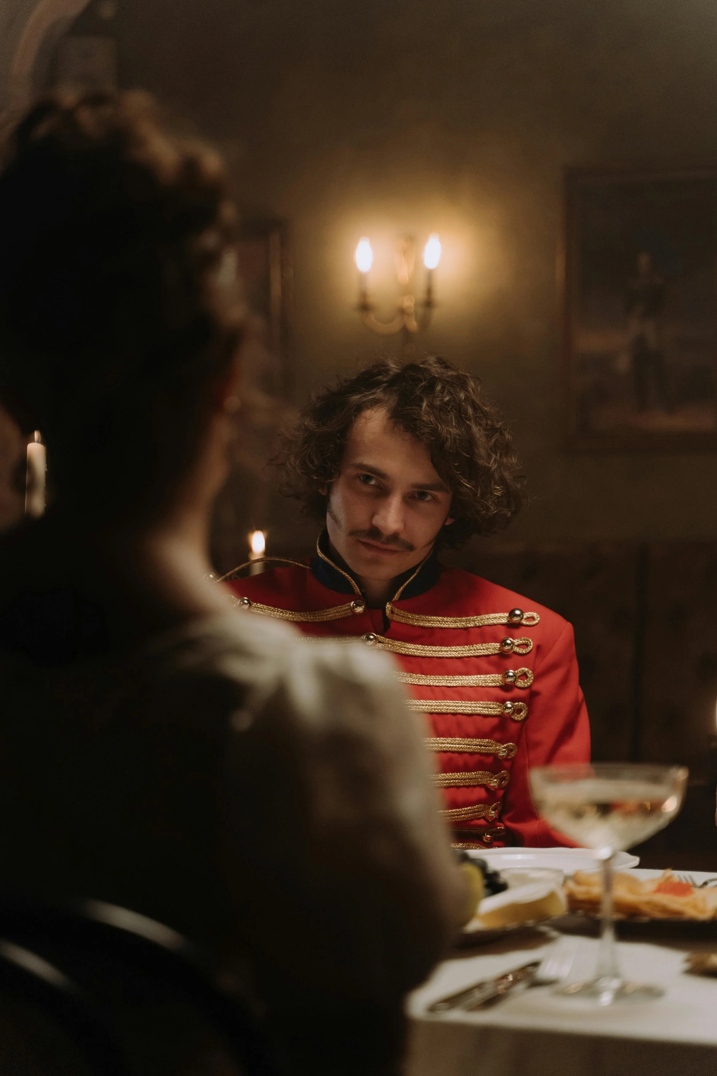A man dressed in a red military-style jacket with gold trim sits at a dinner table, engaged in conversation with a woman whose back is to the camera. The setting appears to be a dimly lit, historic or vintage room with candlelight and portraits on the wall.