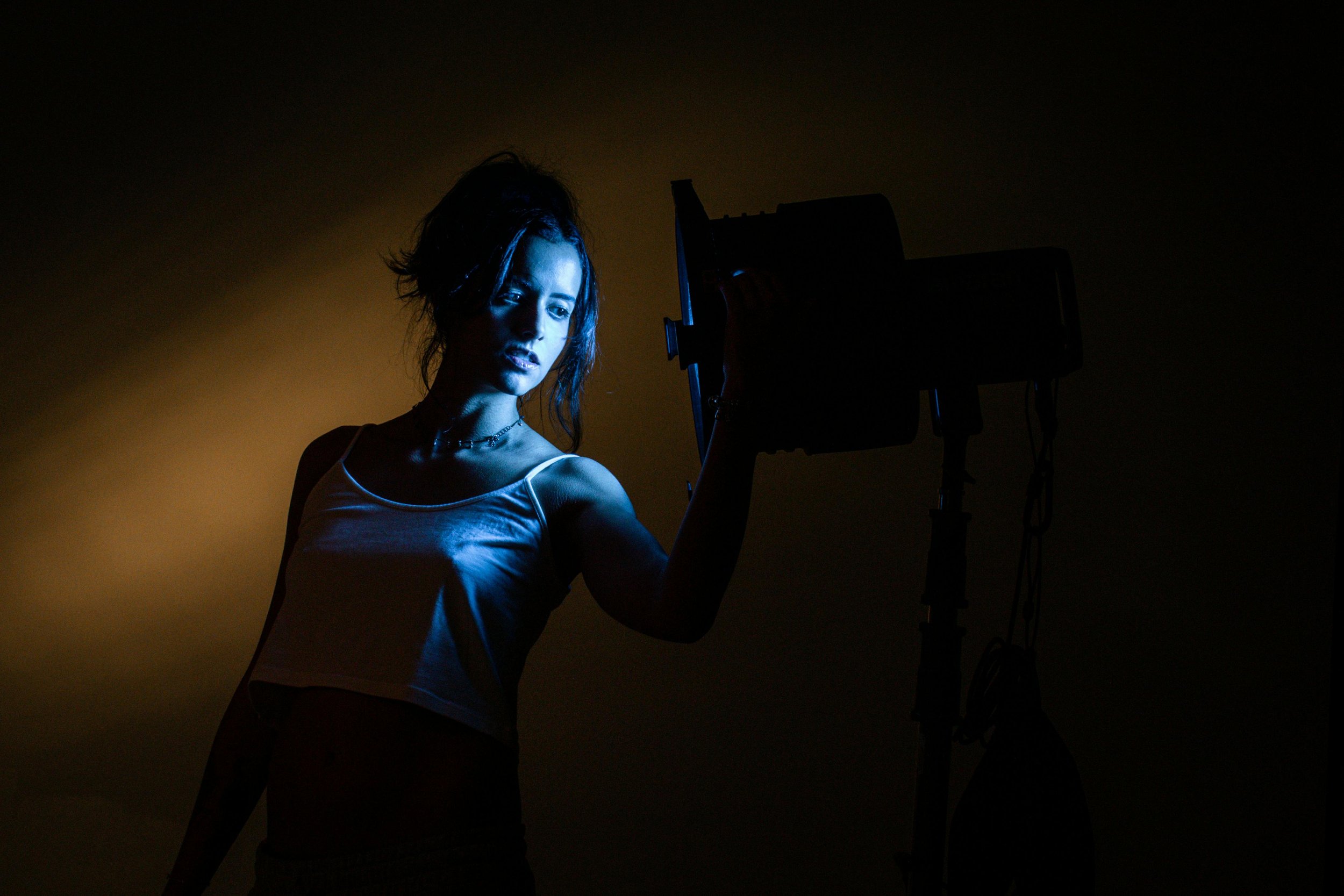 A woman with dark hair, wearing a white tank top, is standing in front of a camera. The scene is dark with a warm light behind her, creating a silhouette effect.