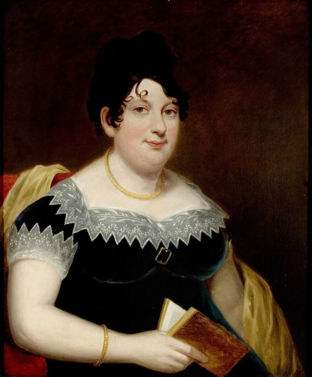 Portrait of a woman with dark hair, wearing a black dress with white embroidery, holding a book, with a gold necklace and bracelet, seated against a dark background.