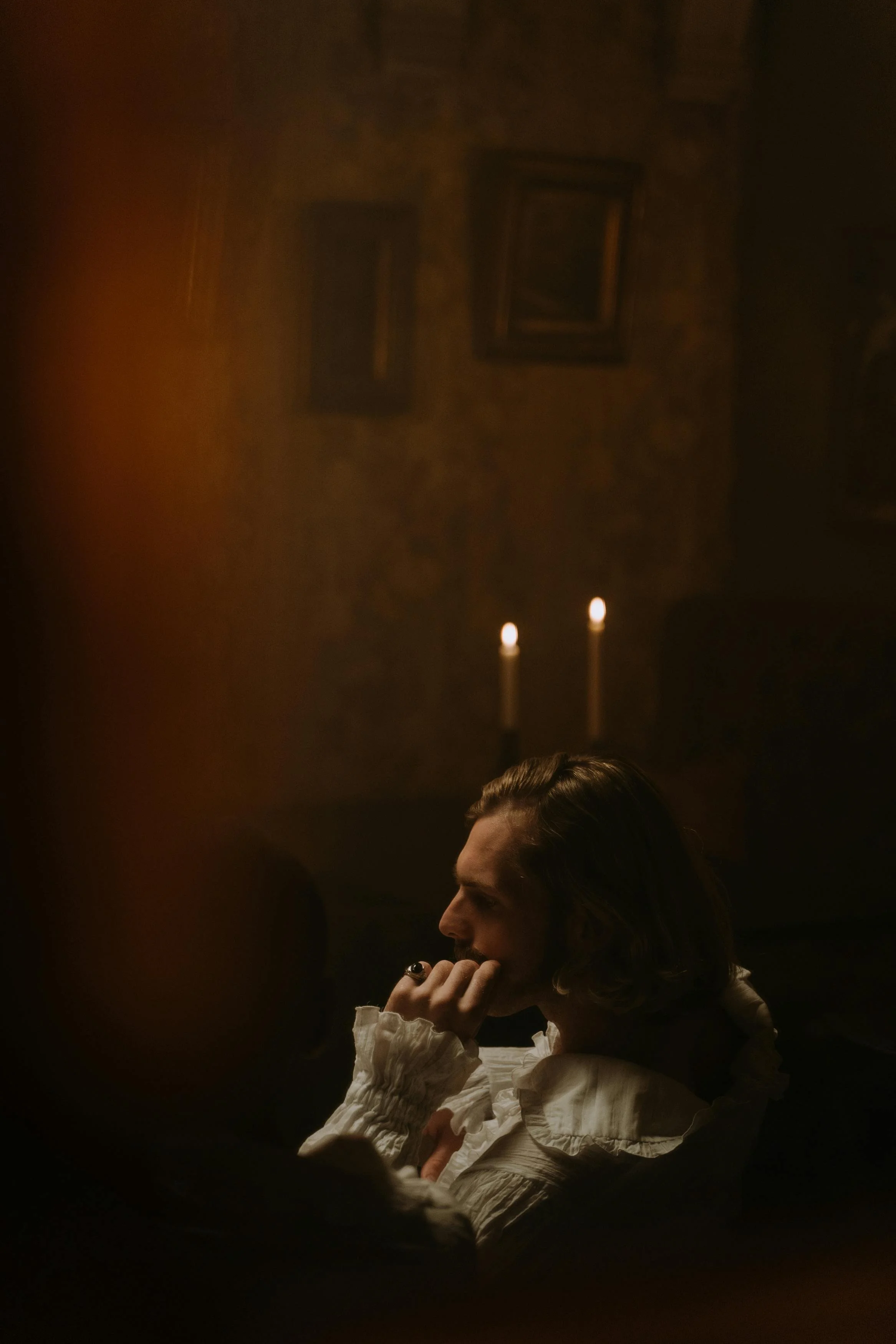 A person with long hair, wearing a white ruffled shirt, sits in a dark room with candlelight, resting their chin on their hand in profile.