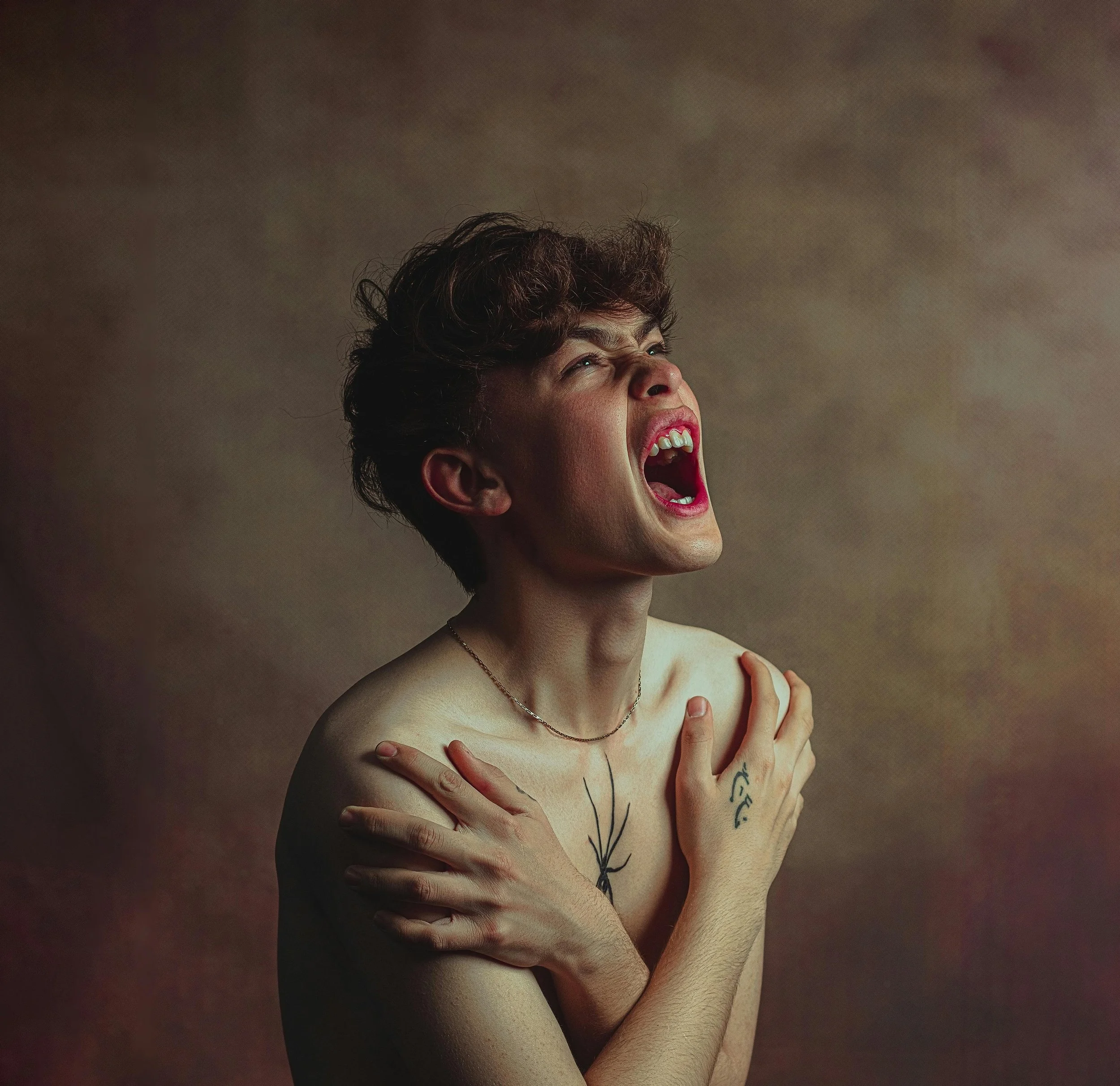 A person with short, curly brown hair is screaming or crying with shoulders crossed over their chest, mouth wide open, eyes closed, and a distressed facial expression. They are wearing a thin necklace and have tattoos on their chest and hand, with a plain, muted background.