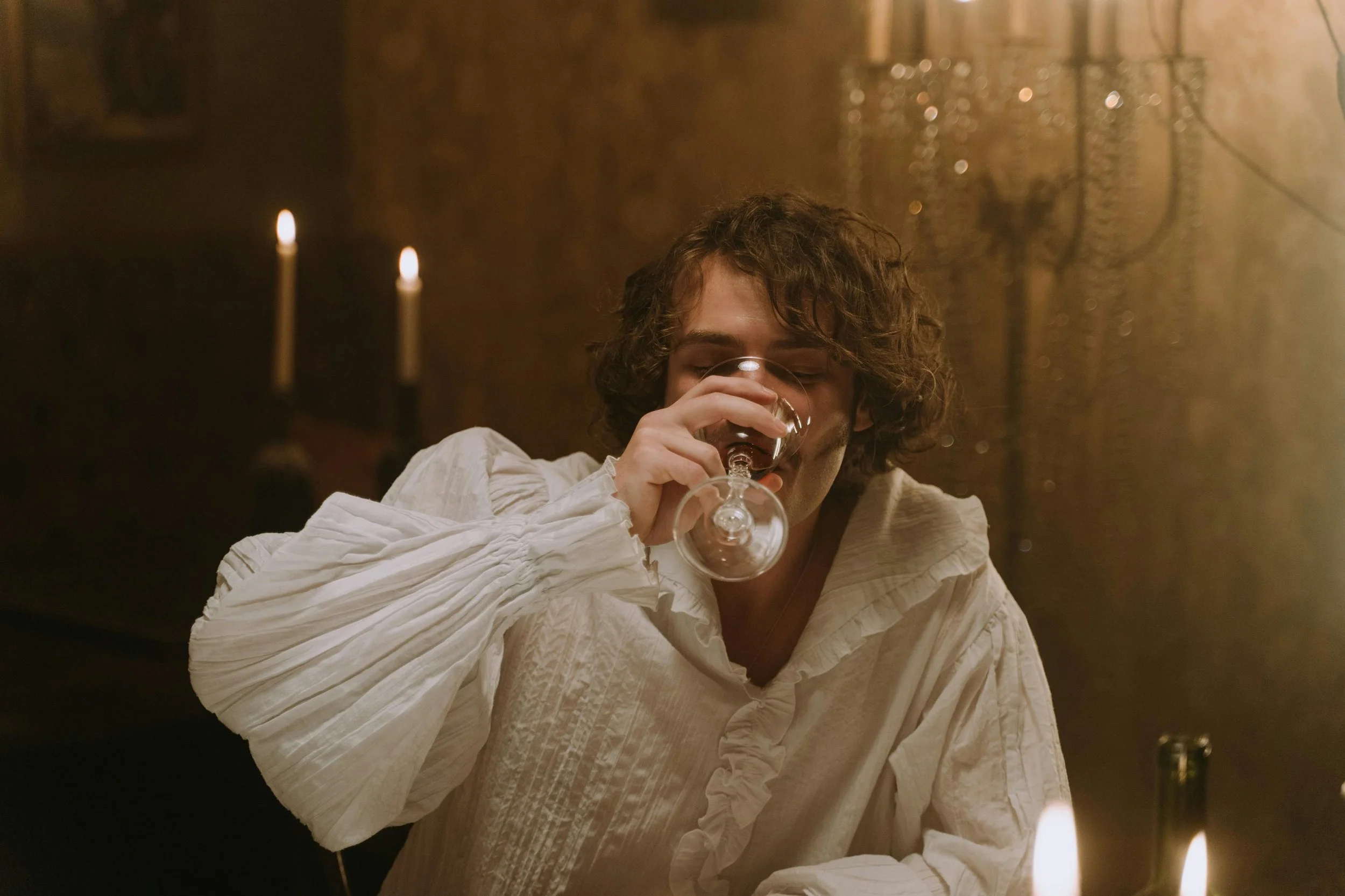 A young man with curly hair dressed in a white vintage-style shirt, drinking wine from a glass in a dimly lit room with candles and ornate decorations in the background.