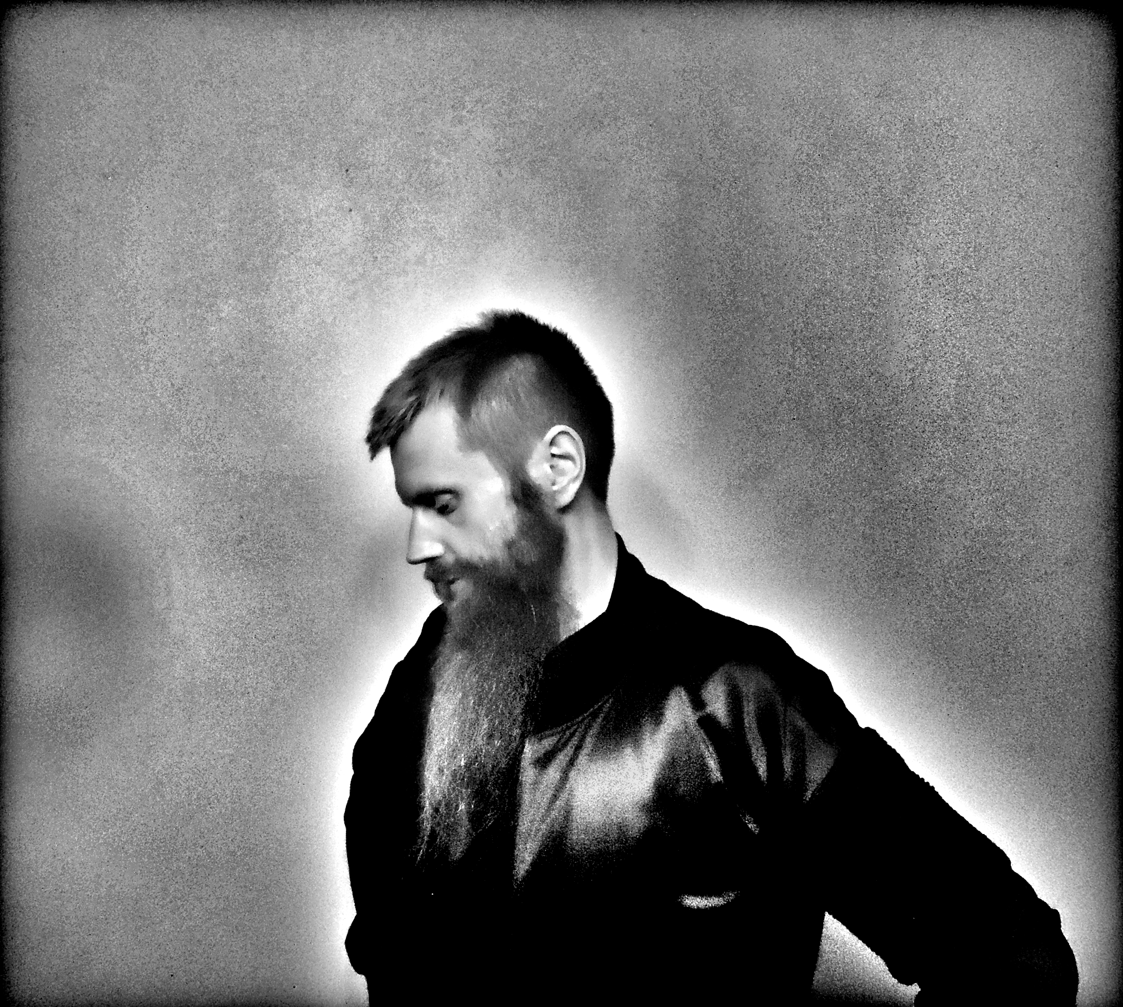Black and white photograph of a man with a beard and short hair, looking down with a halo-like glow around his head and shoulder.