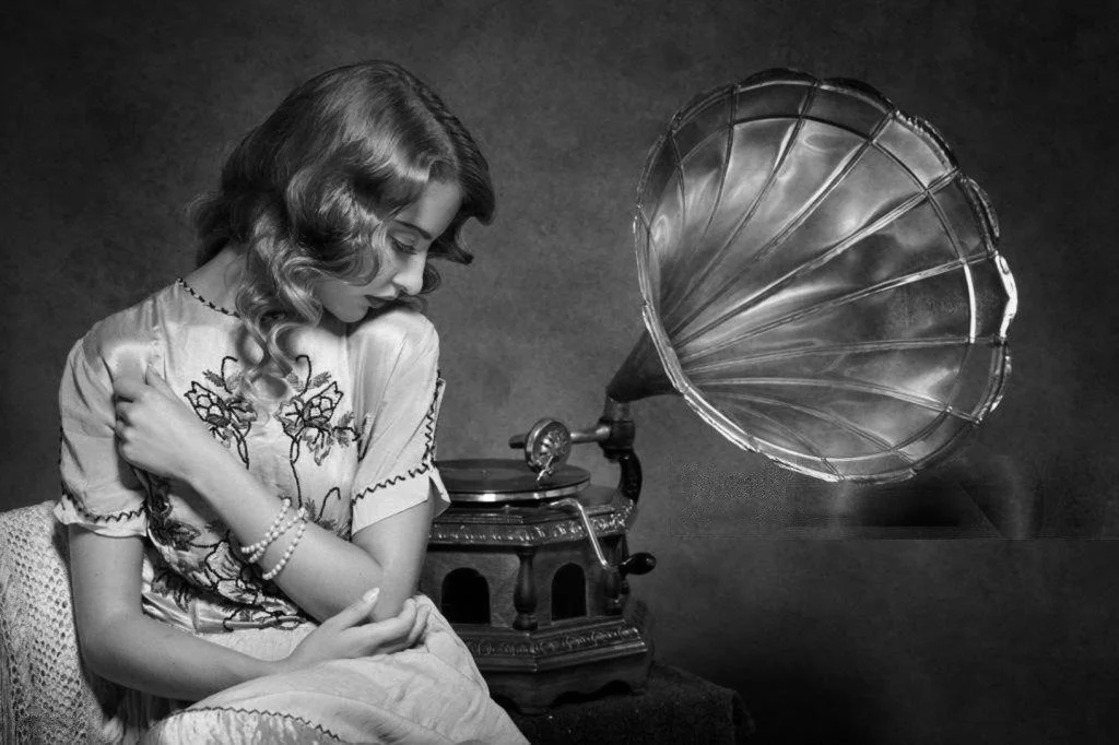 A woman with wavy hair, wearing a floral dress and pearl bracelet, sitting next to an antique gramophone in a black and white photo.
