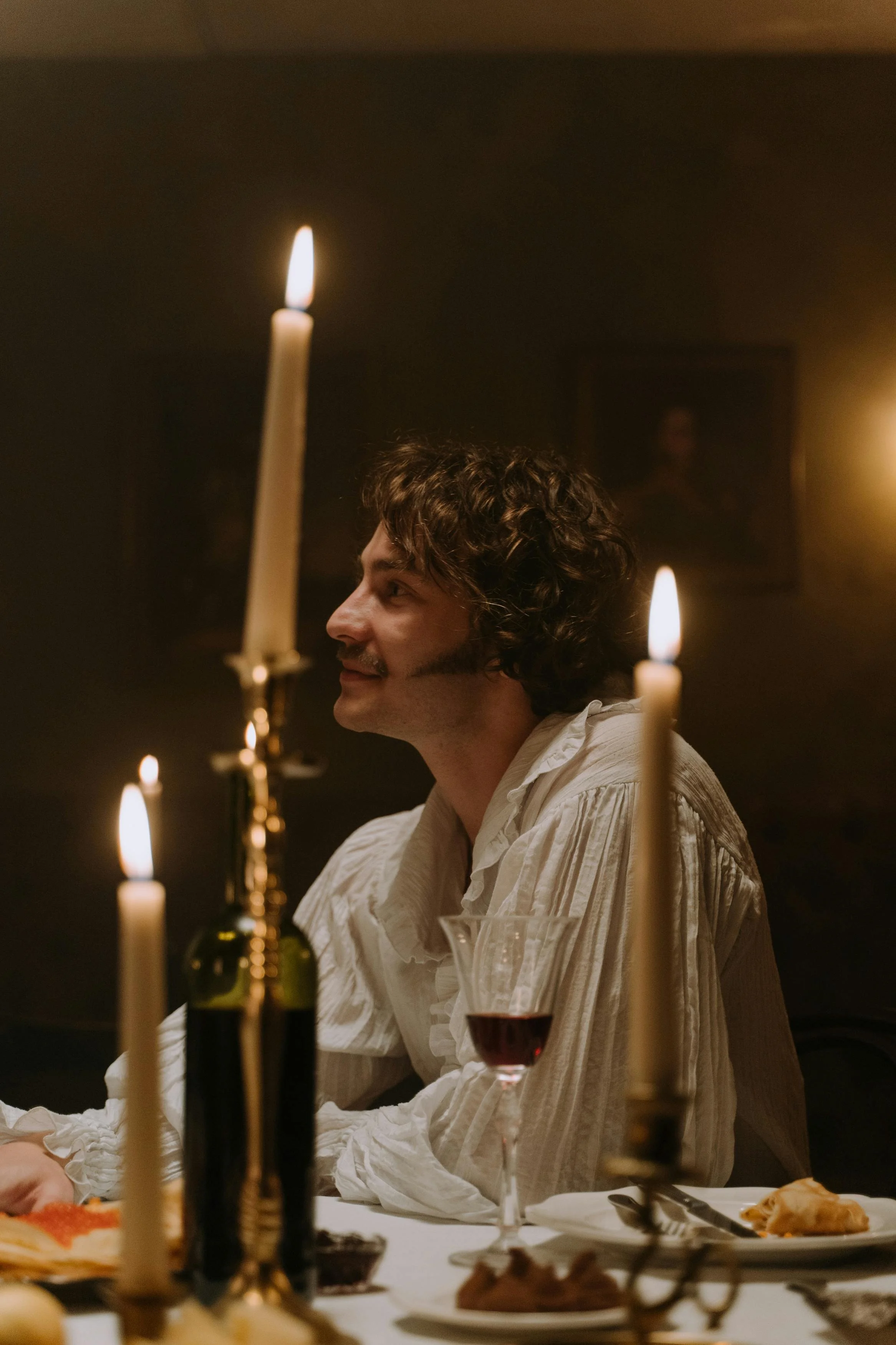 A man with curly hair and facial hair is sitting at a dining table, smiling. The scene is dimly lit with candles, and there is a glass of red wine in front of him. The background has a dark wall with a picture frame.
