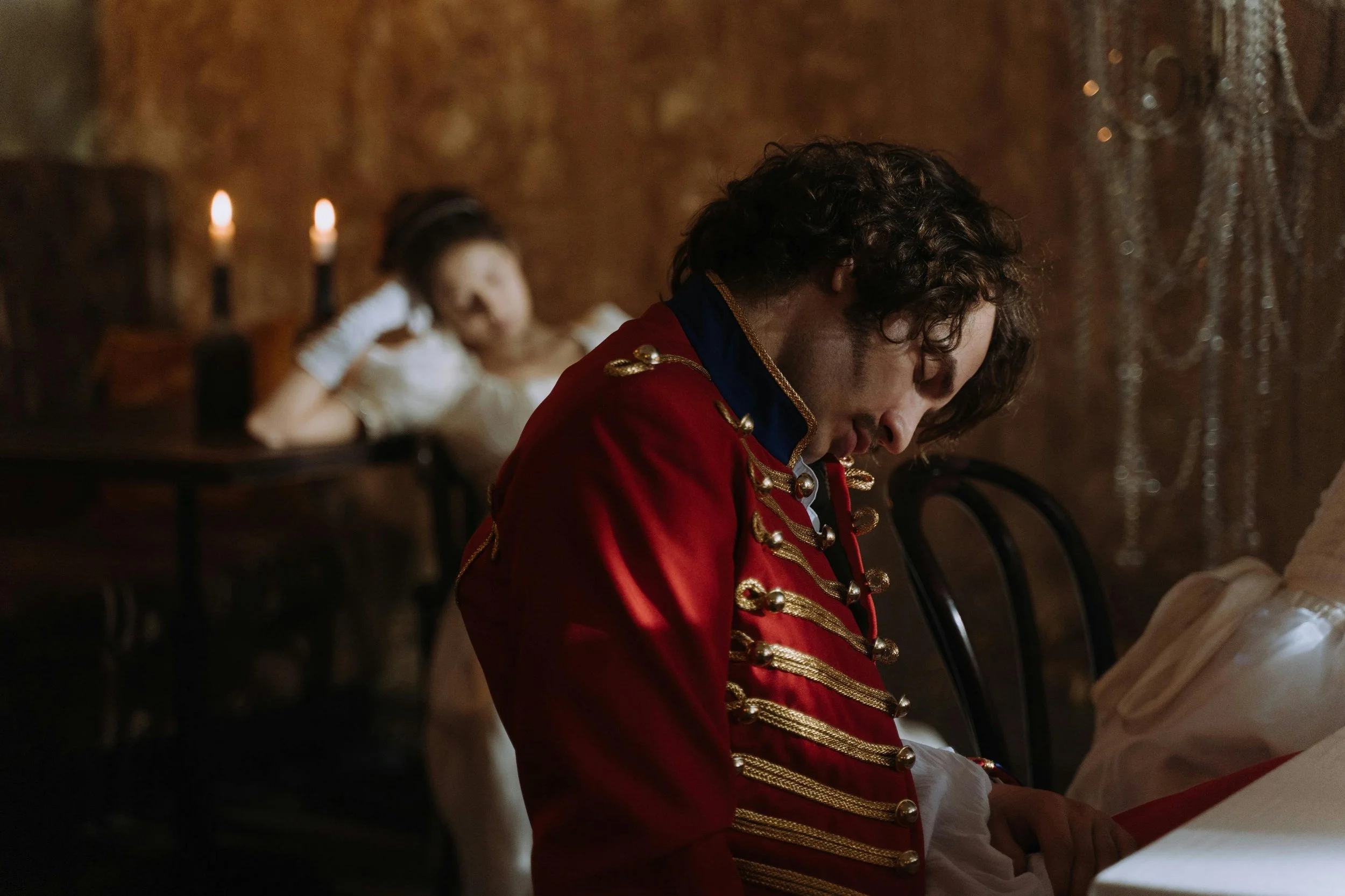 A young man dressed in a red military-style jacket with gold details is sitting at a table with his head bowed. A girl in a white dress is lounging on a sofa in the background, resting her head on her hand.