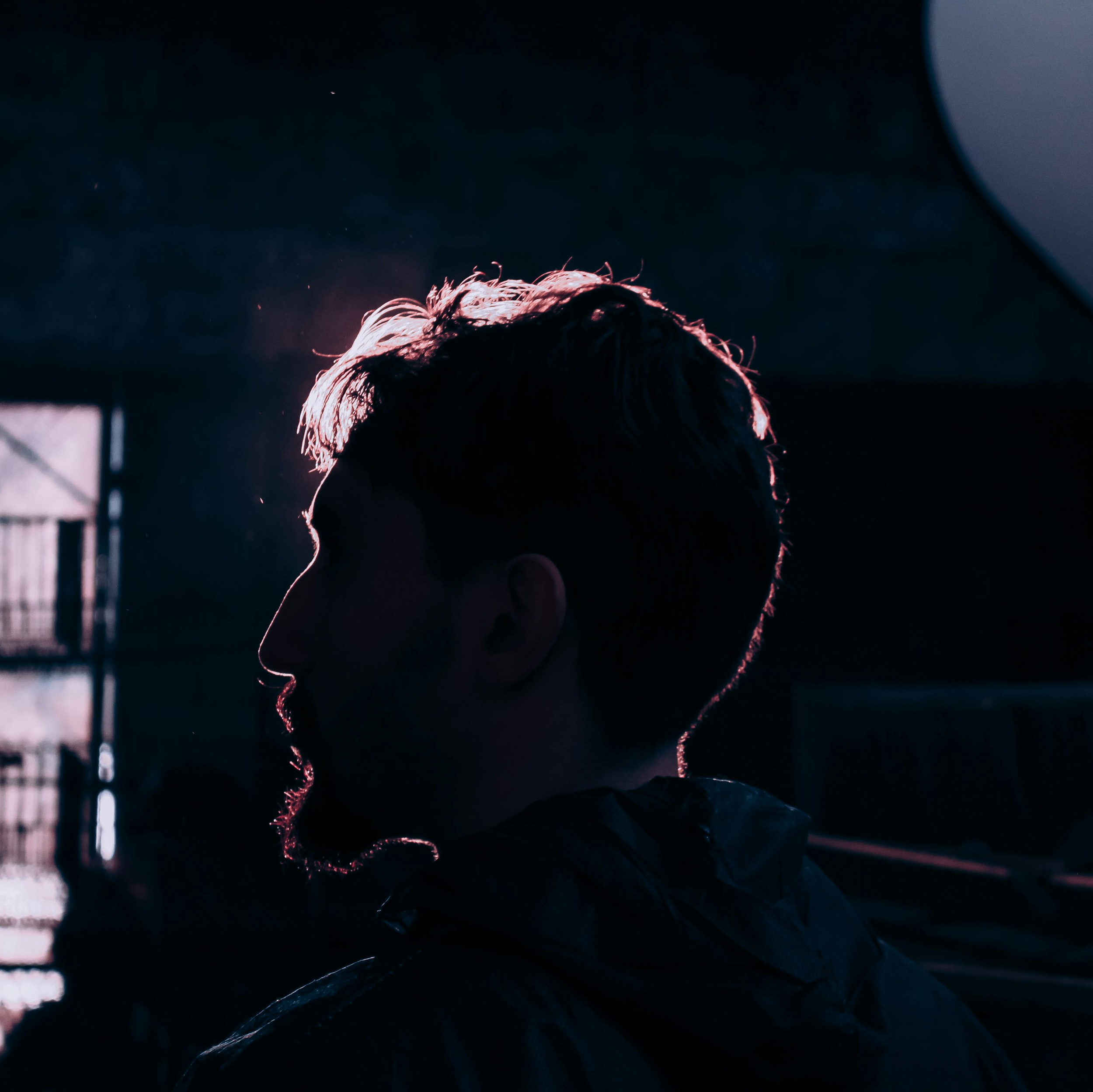 Silhouette of a person with styled hair, backlit by sunlight, in an urban setting with a fence and building structure in the background during sunset or sunrise.